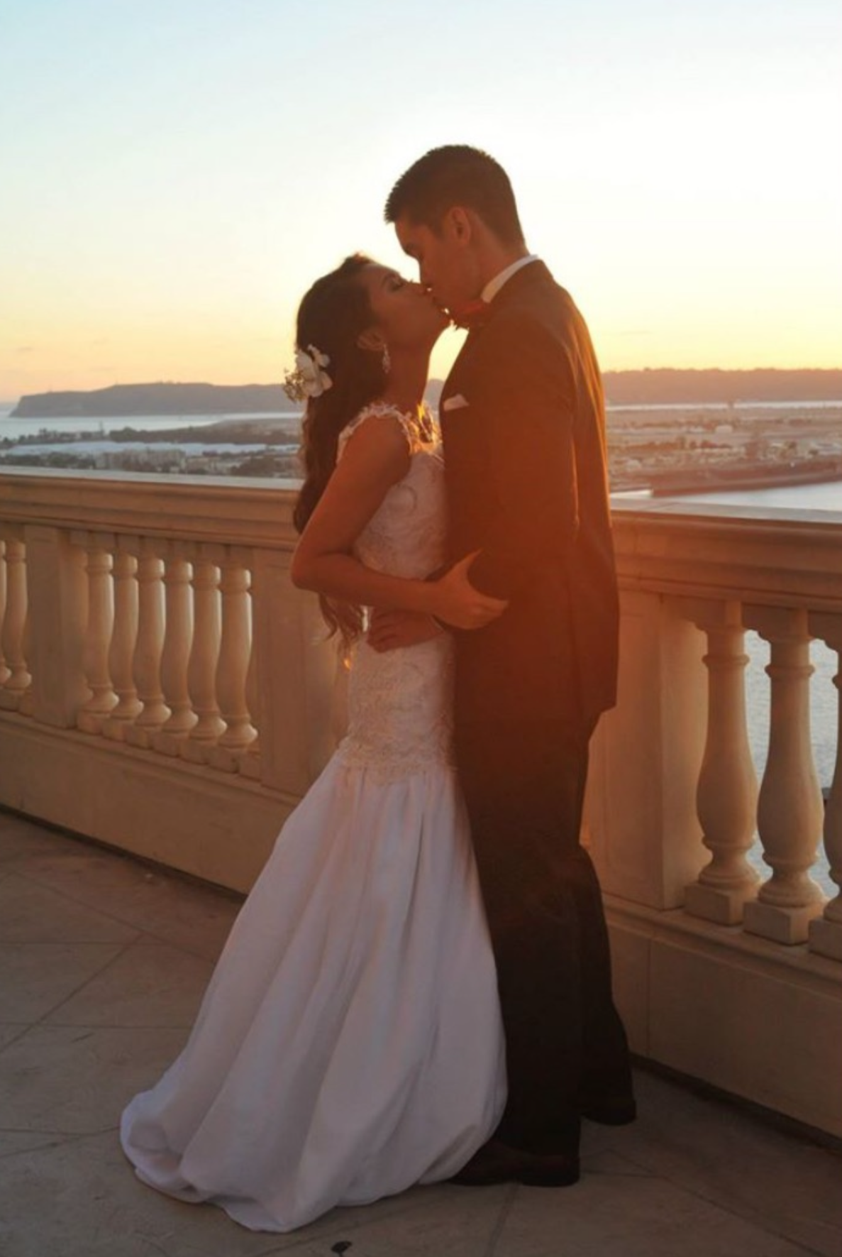 A bride and groom share a kiss on a balcony during sunset, with a view of water and distant land in the background.