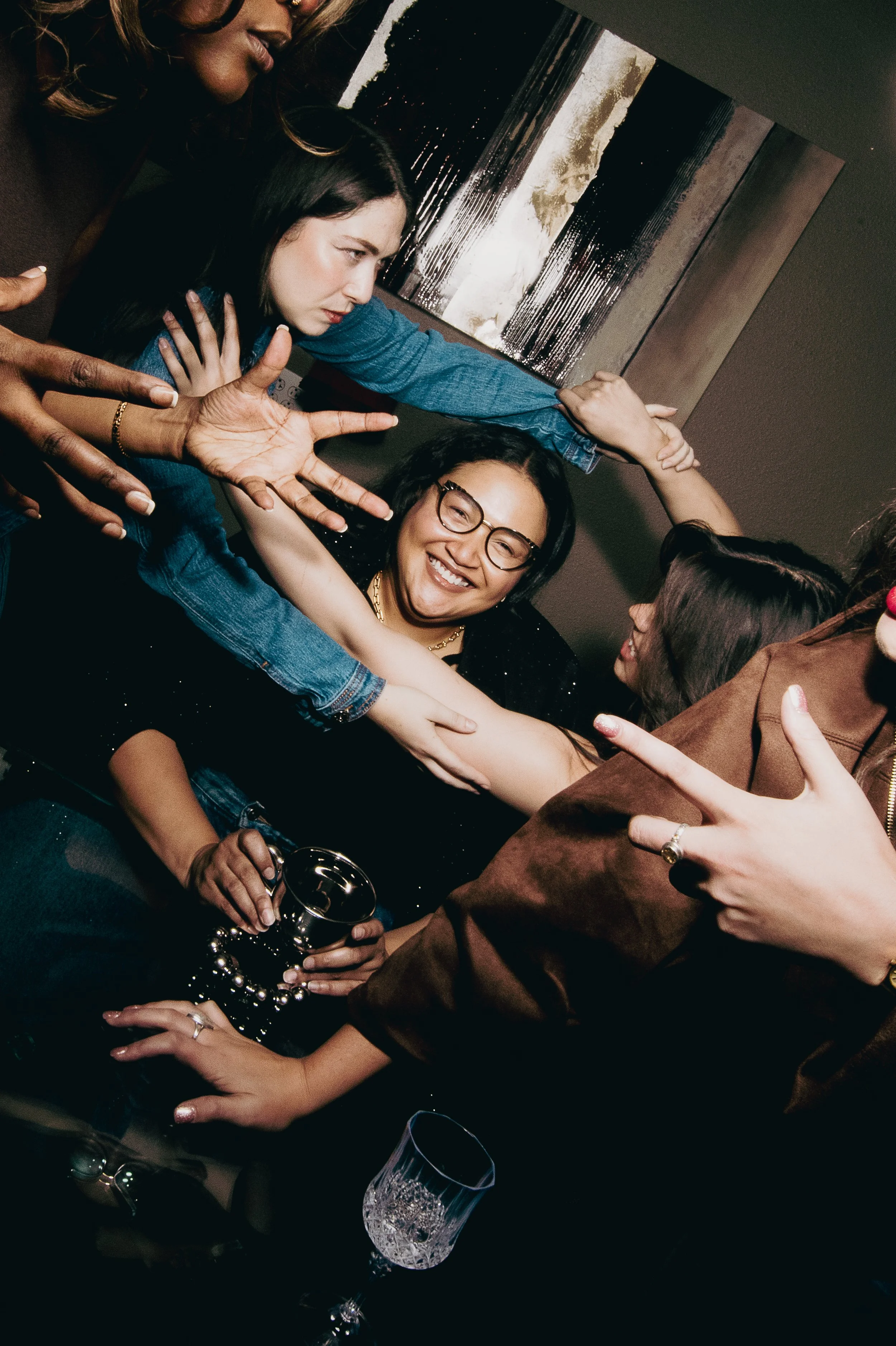 A group of diverse women sitting closely together, smiling, laughing, and engaging in a lively, physical group hug at a social gathering. One woman in the center is holding a glass of champagne, and others are touching each other affectionately. There is a modern abstract painting on the wall behind them.