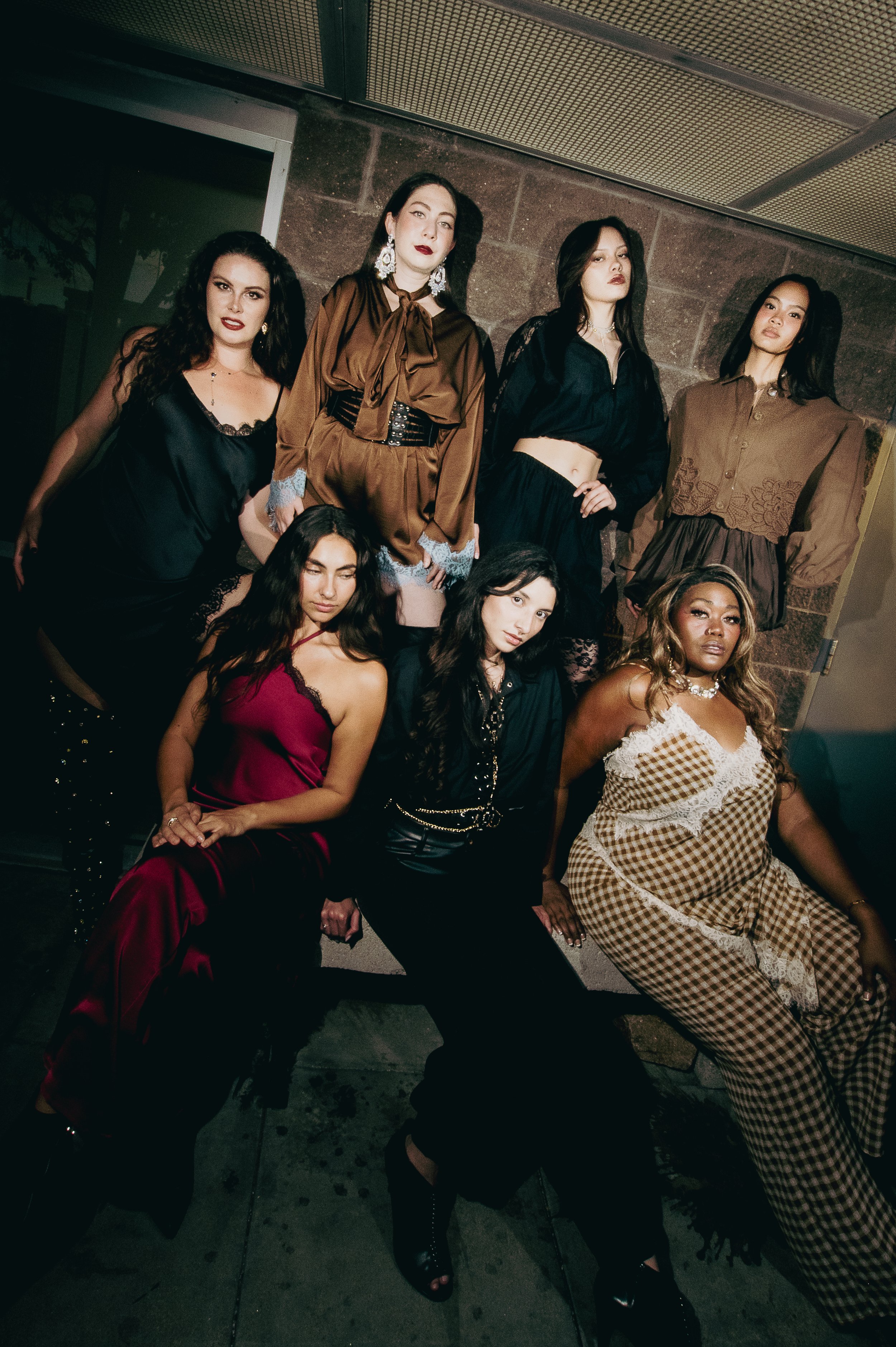 A group of seven women with diverse appearances and styles posing together against a brick wall and a metal ceiling. The women are standing or sitting in various outfits, including satin, lace, leather, and plaid, with some wearing jewelry and makeup. The setting appears to be indoors or on a city street at night.