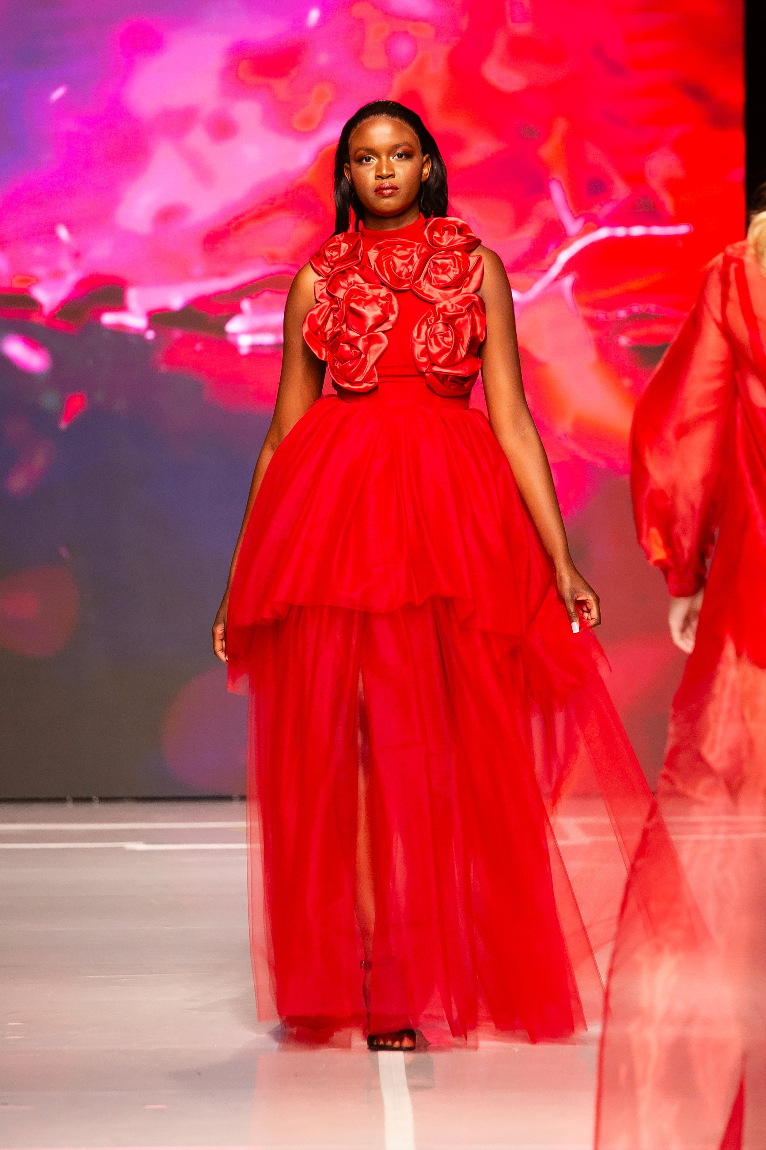 A model walking on a runway wearing a red dress with floral embellishments on the bodice and a flowing tulle skirt, with a colorful abstract background.