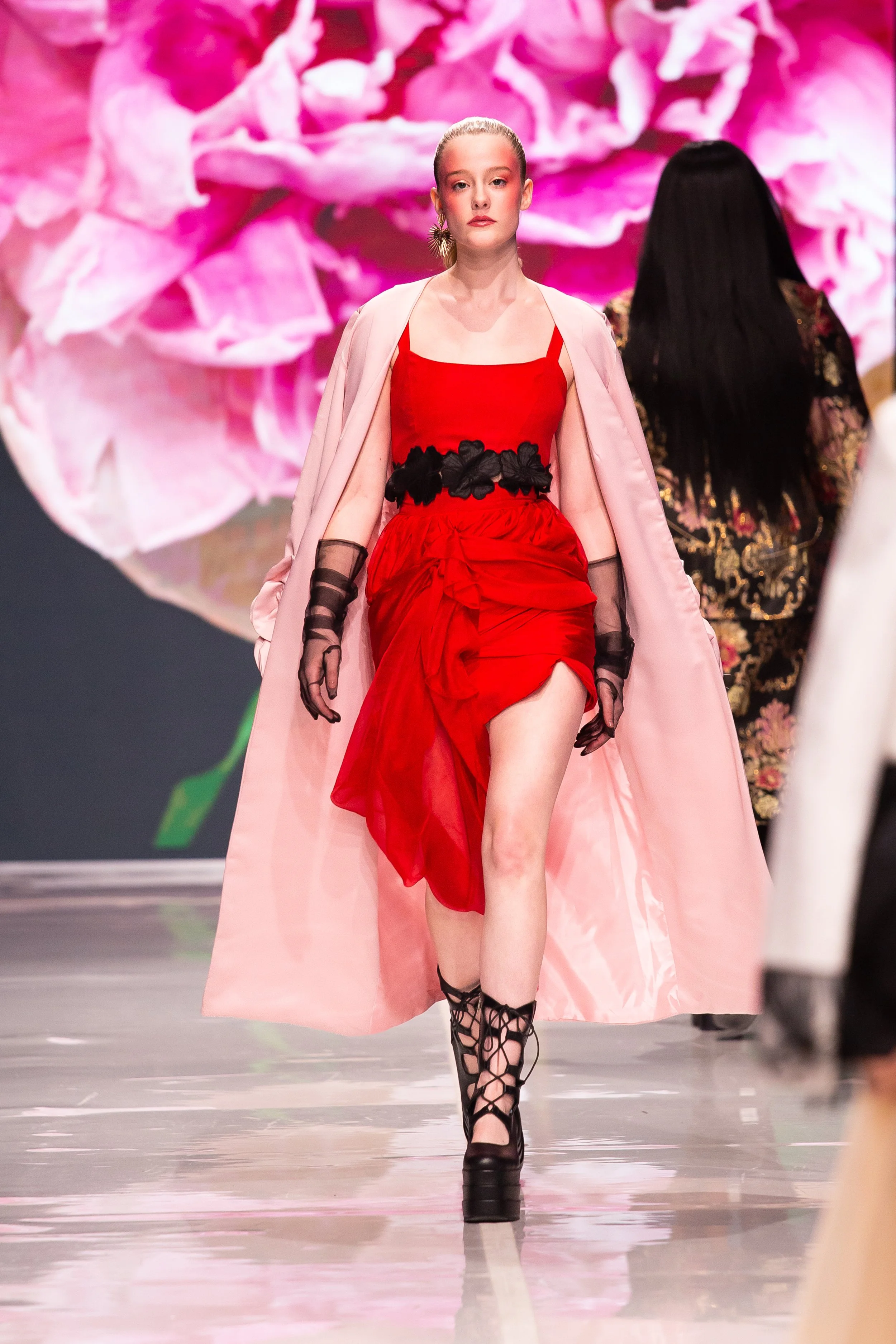 Model walking on runway wearing a red dress with black floral accents, sheer black gloves, and black lace-up platform shoes, with a pink cape and a large floral backdrop.