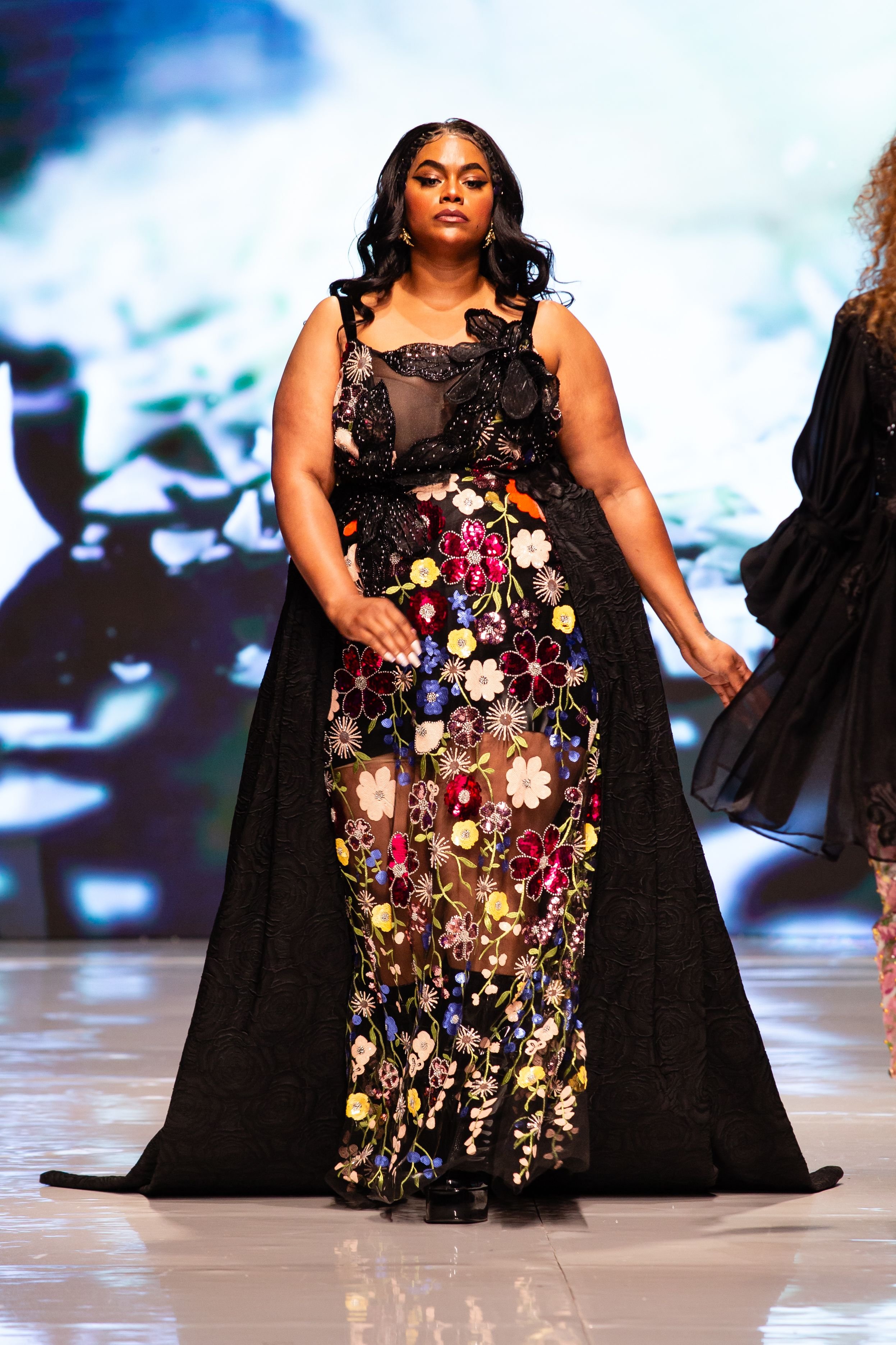 A woman with dark hair and earrings walking on a stage, wearing a black and floral embroidered gown with a long black train.