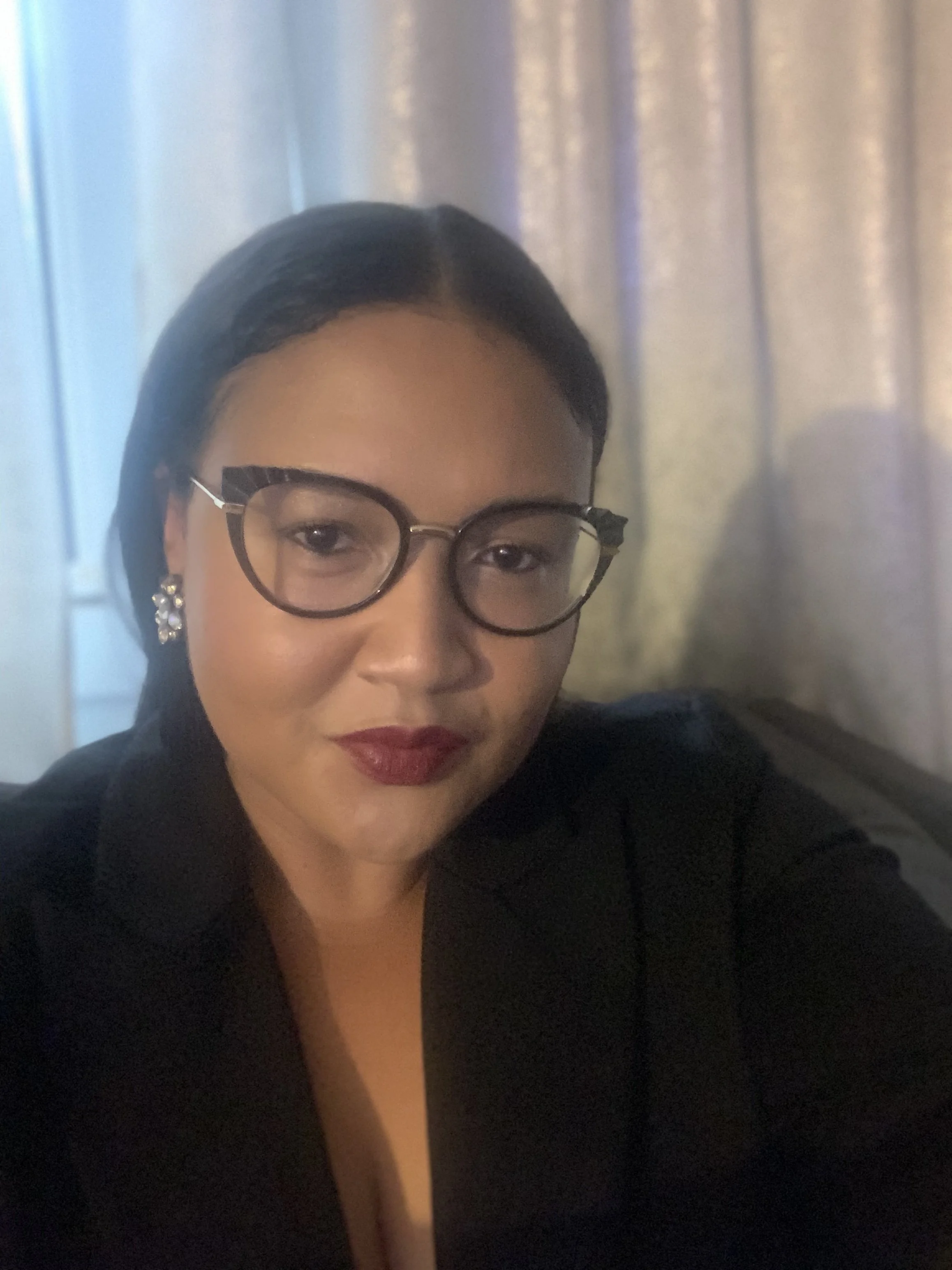 Close-up of a woman wearing glasses and earrings, with makeup, in front of a blurred background.