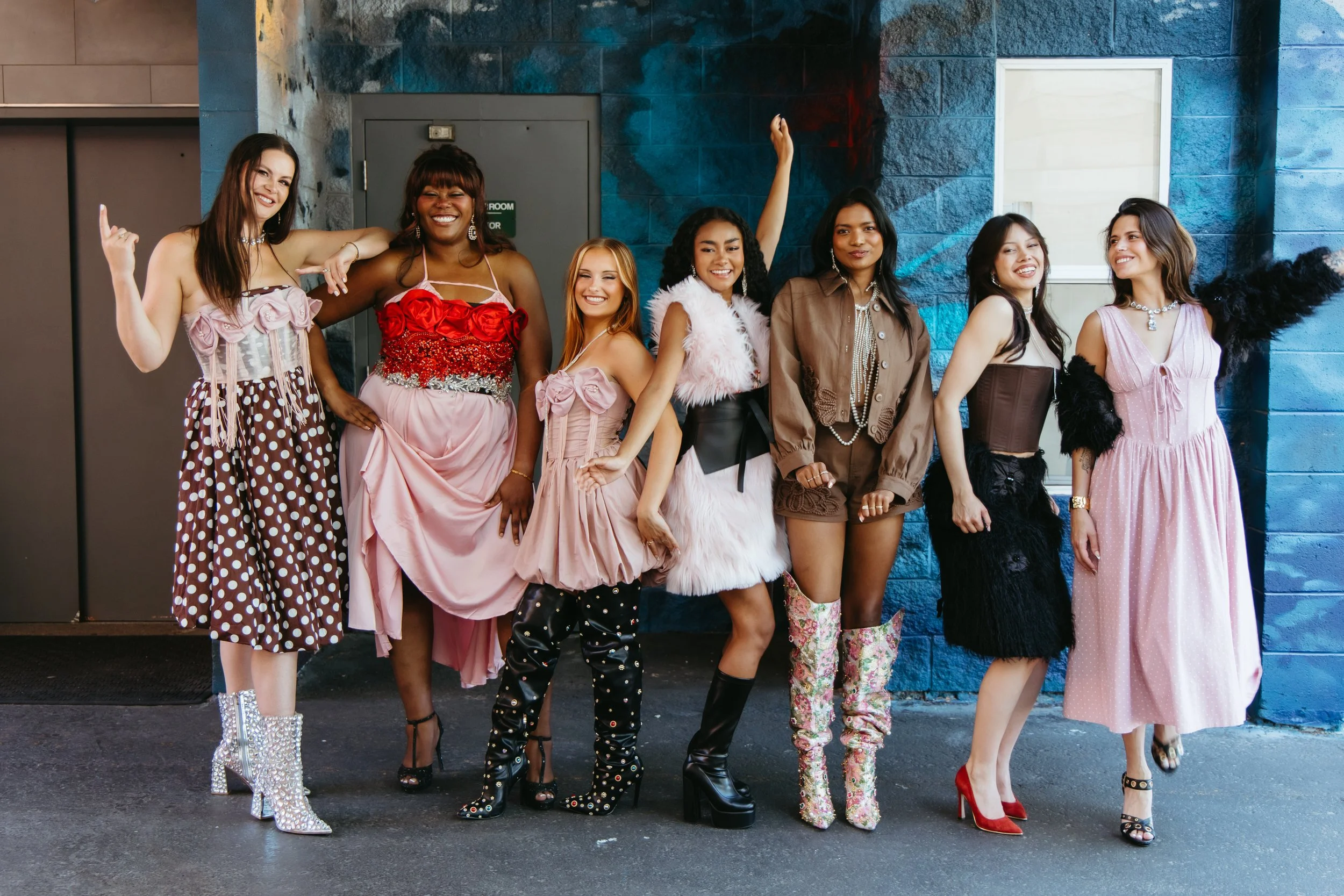 Group of women dressed in fashionable and glamorous clothing, standing together outside against a blue textured wall, smiling and posing for the camera.