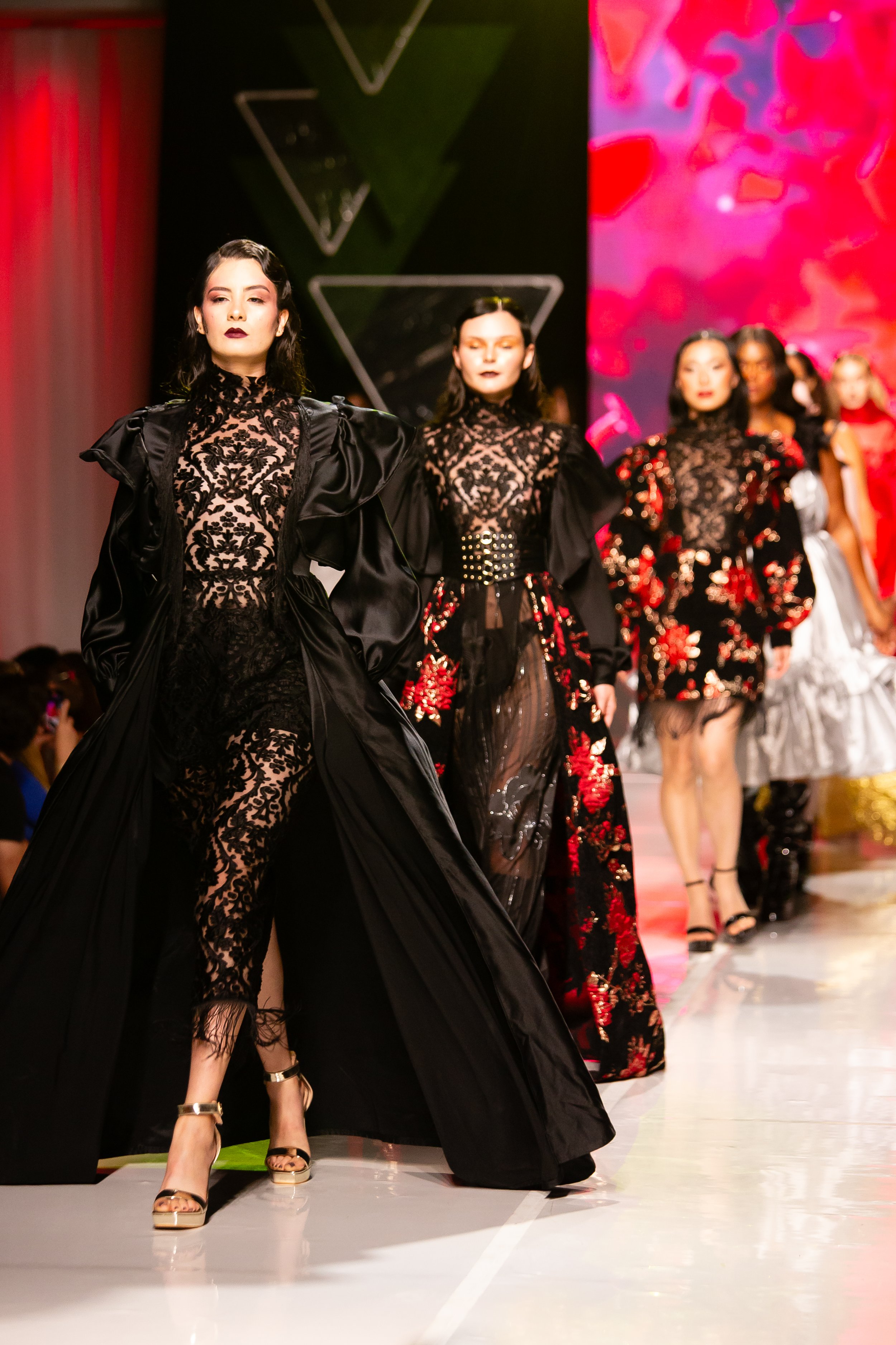 Models walking runway in black lace and floral dresses at a fashion show.
