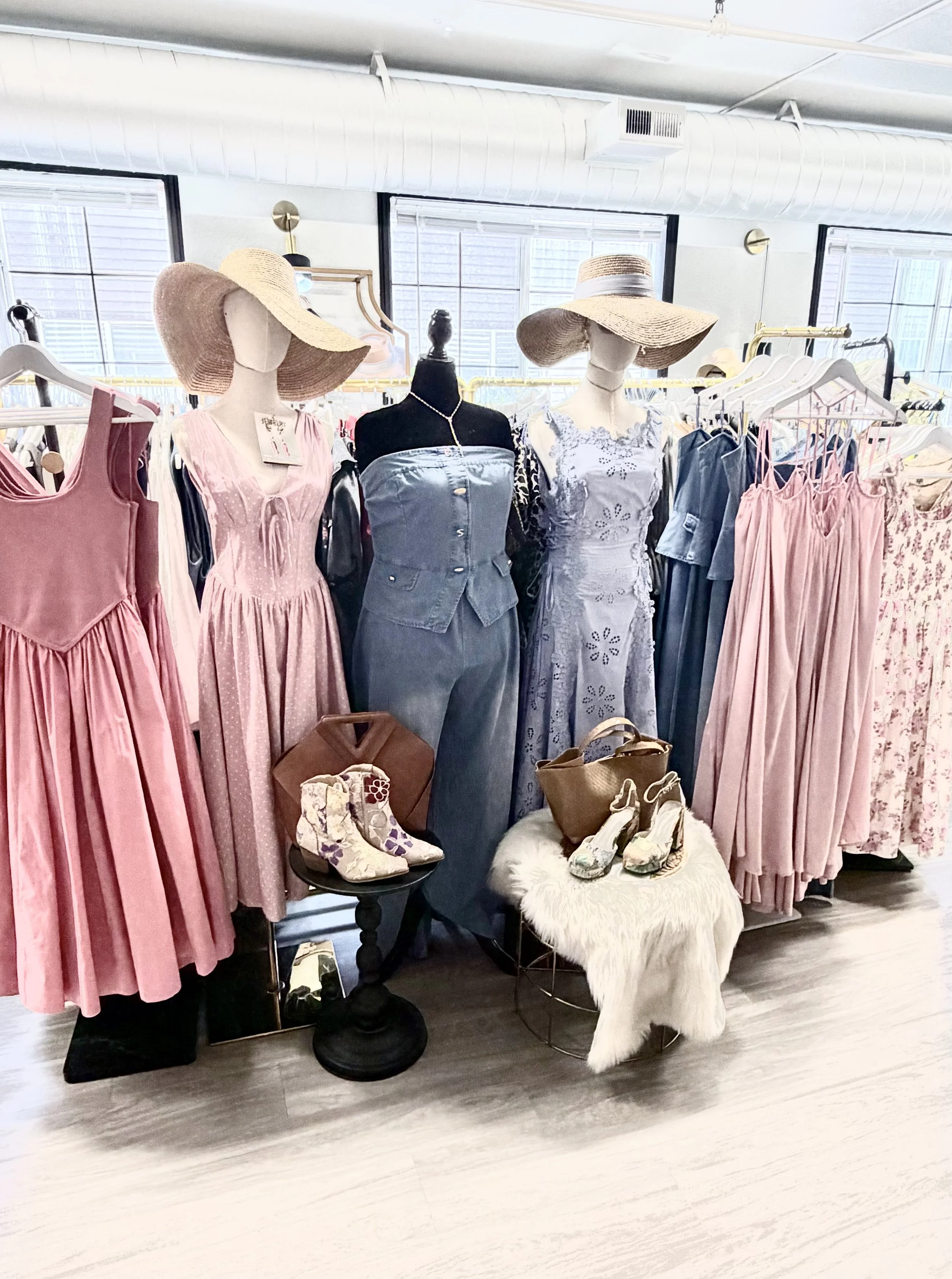 Display of women's clothing and accessories in a boutique, featuring mannequins dressed in pastel dresses and wide-brimmed hats, with items on hangers and a small table with shoes and bags.
