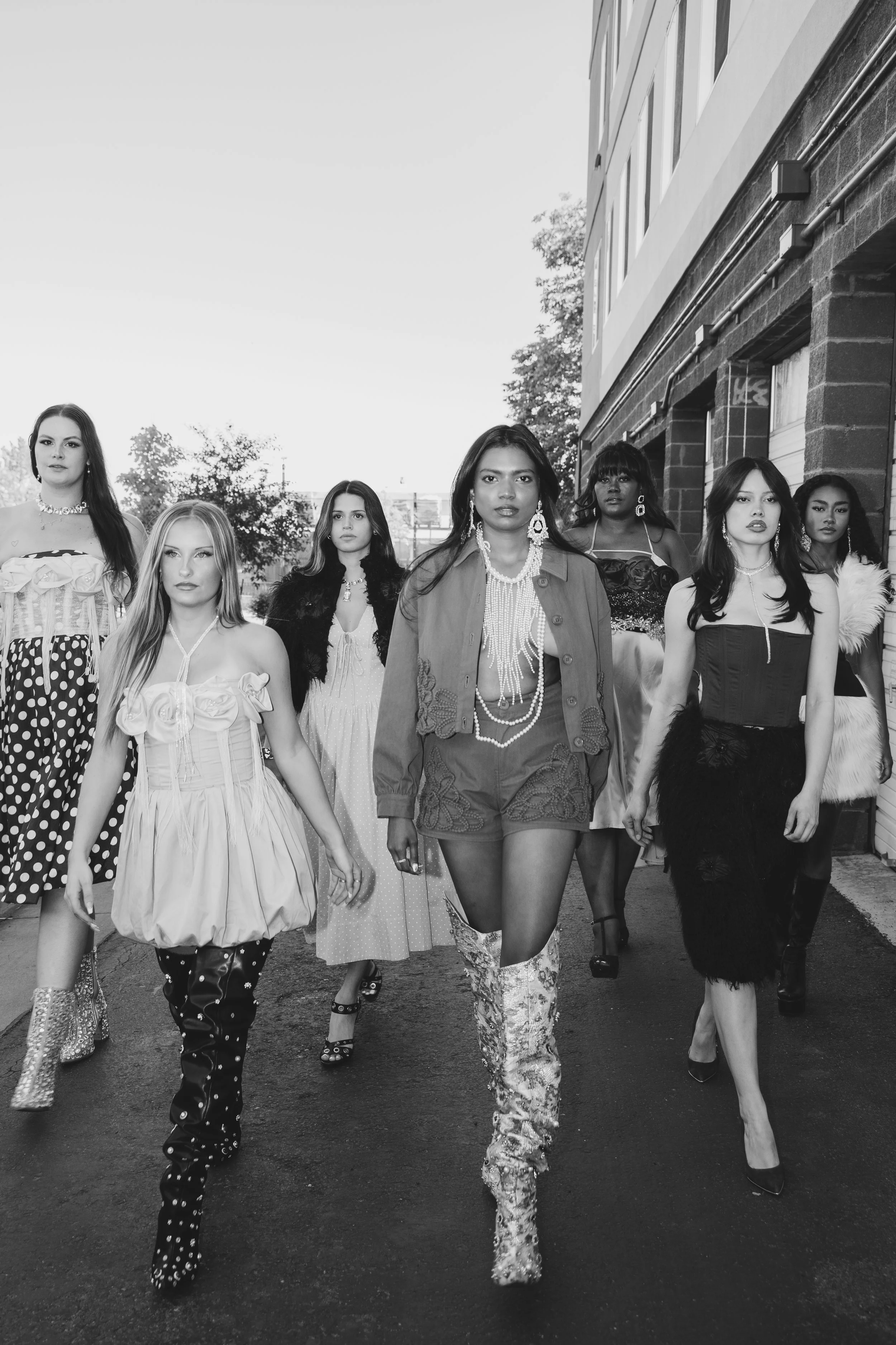 A group of seven diverse women walking on a street, dressed in stylish and fashionable clothing, with some wearing jewelry and high heels, in an urban setting with a brick building and trees in the background.