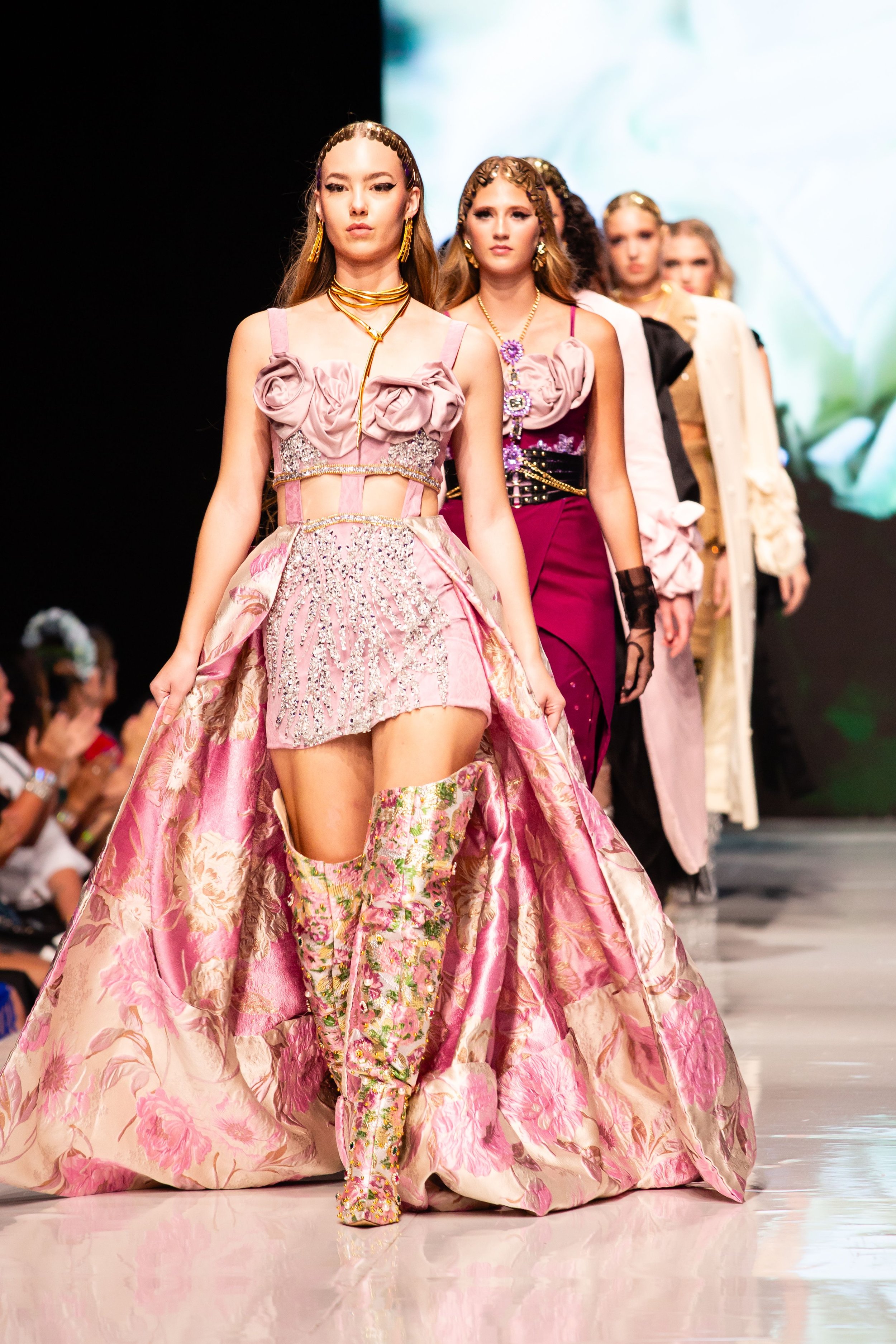 Models walk down the runway during a fashion show, wearing elaborate pink and floral couture dresses and matching accessories.