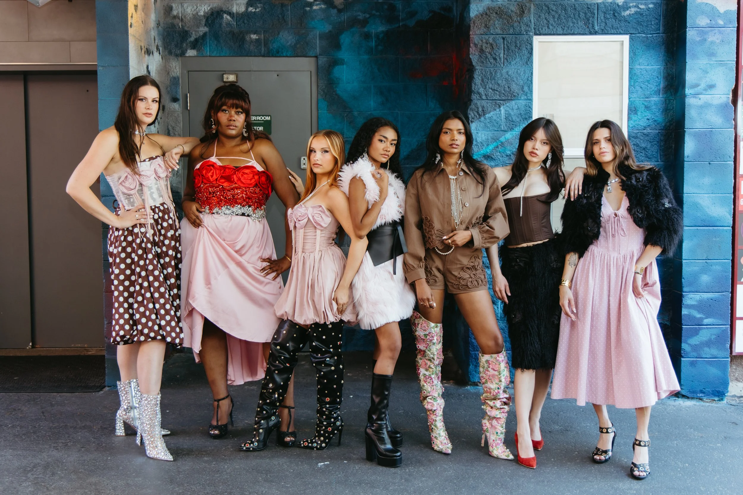 Seven young women standing together against a blue wall, dressed in fashionable and colorful outfits.