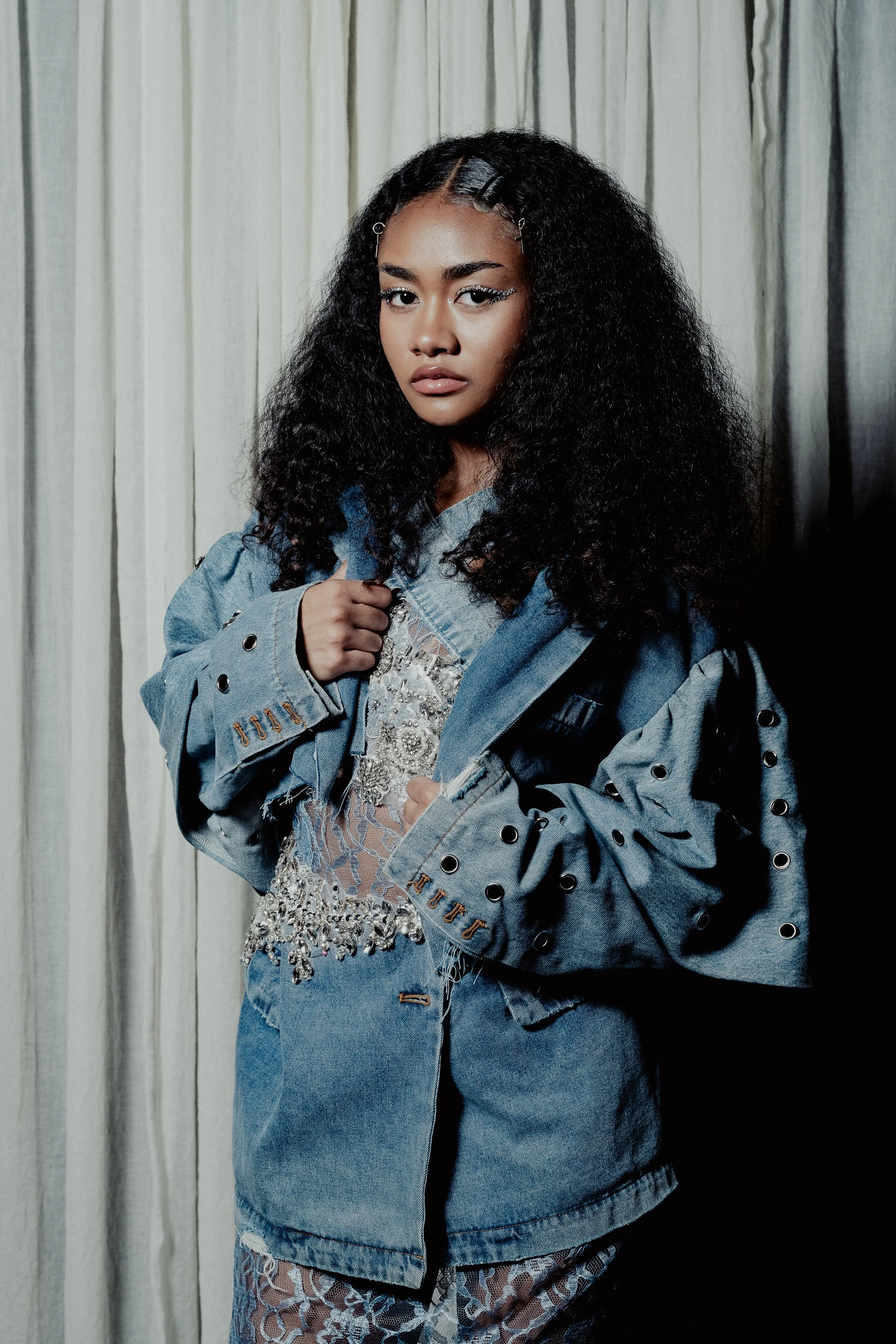 A young woman with curly black hair and hair clips, wearing a distressed oversized denim jacket and lace clothing, standing in front of a curtain background.