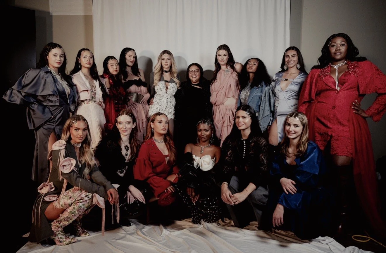 Group of women posing together in fashionable outfits, some sitting and some standing, in front of a plain backdrop at an indoor event.