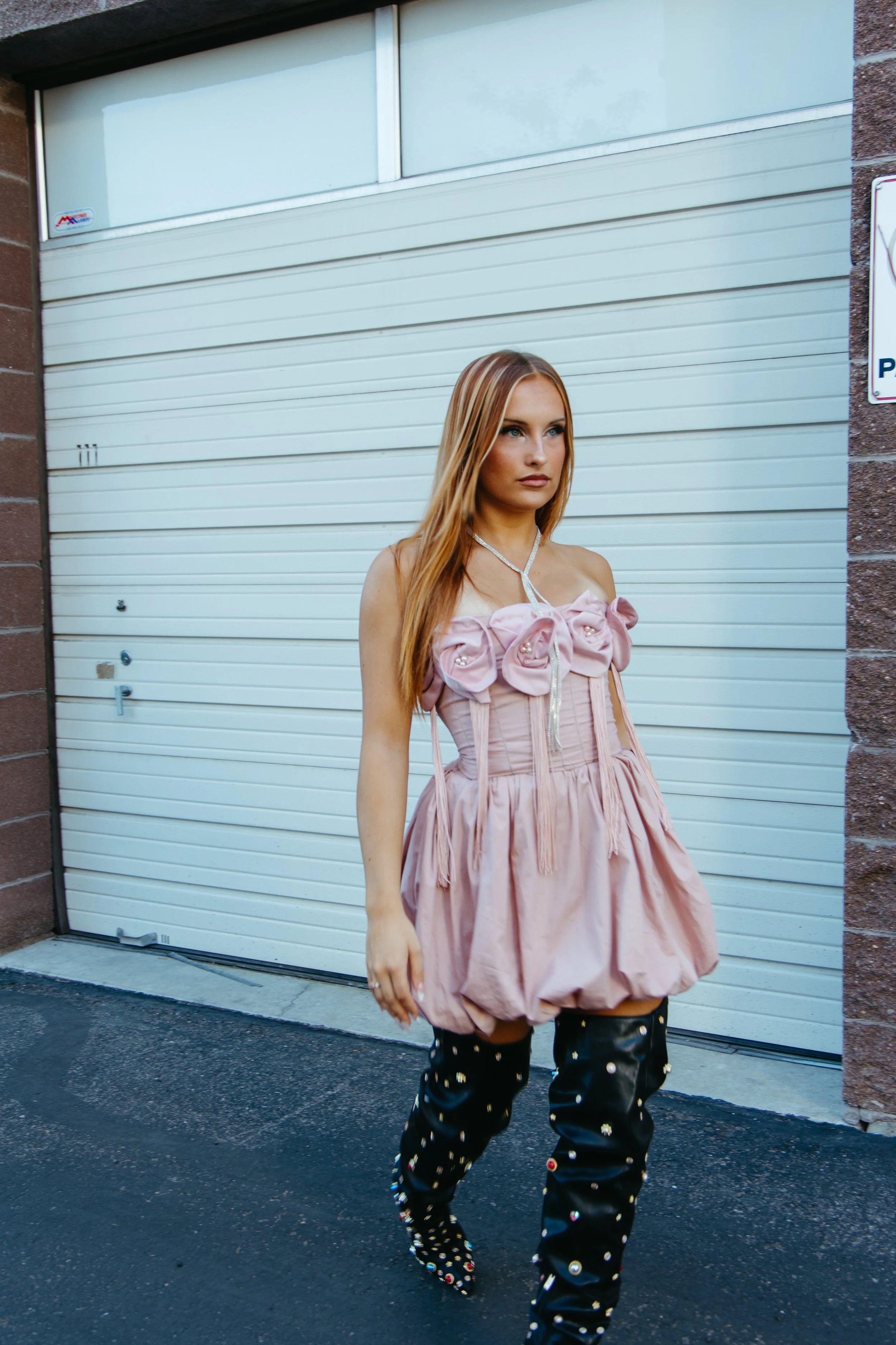 A woman standing in front of a white garage door with a brick wall on either side, wearing a pink strapless dress with large fabric flowers, and black thigh-high boots decorated with multicolored rhinestones.