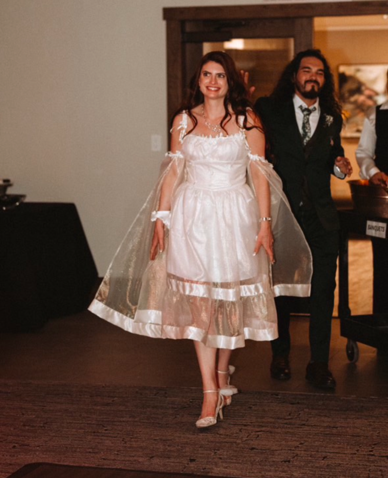 Woman in a white wedding dress standing indoors with a man in a black suit behind her, smiling.