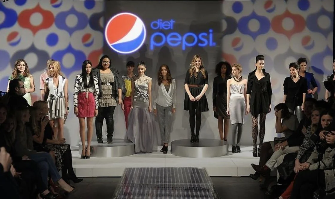 A group of women showcasing fashion on a runway during a Pepsi Diet event, with an audience seated on either side.