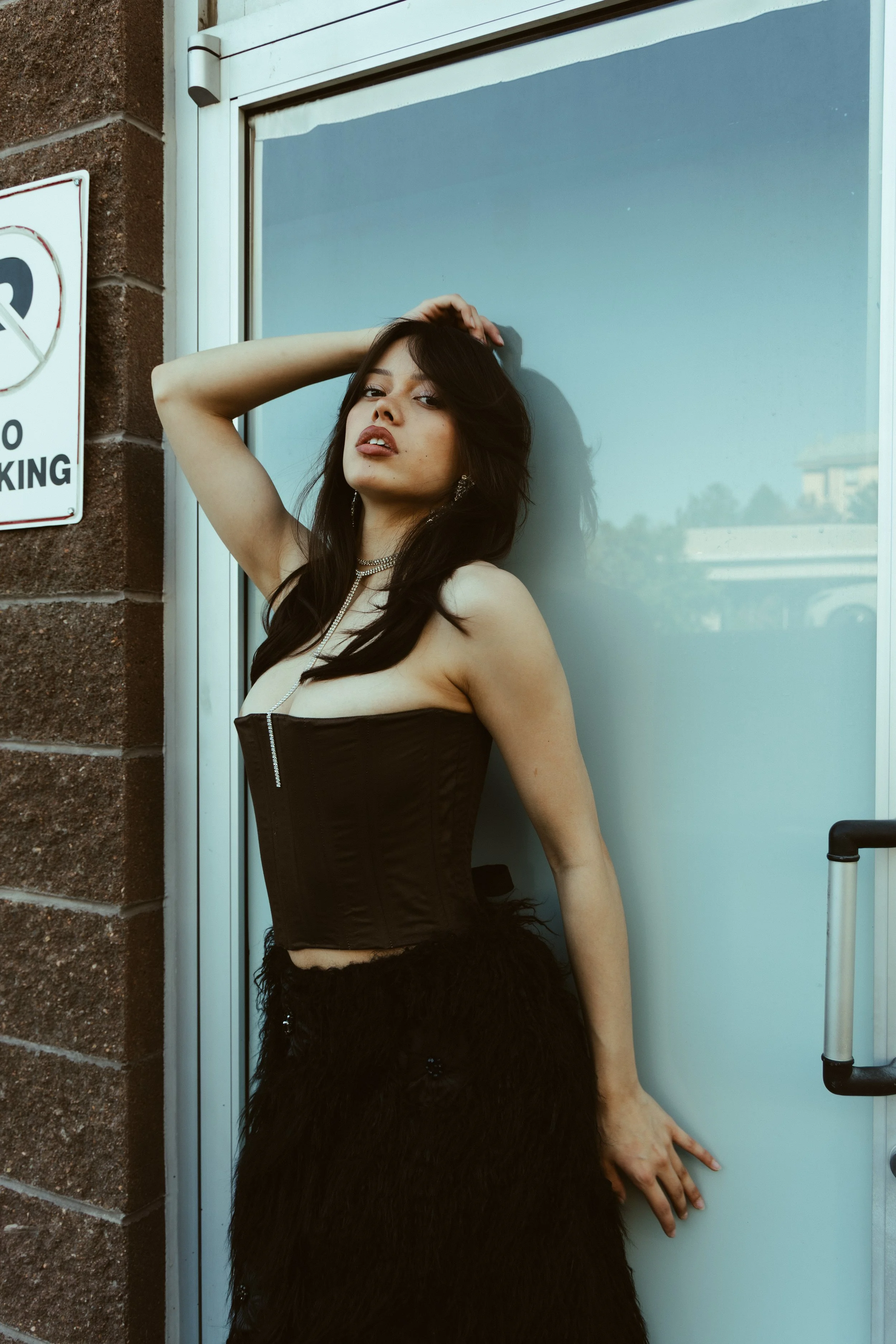A woman dressed in black lingerie and a fluffy black skirt leaning against a glass door, with her arm bent behind her head and her hand resting on her hair, posing outdoors near a brick wall and a