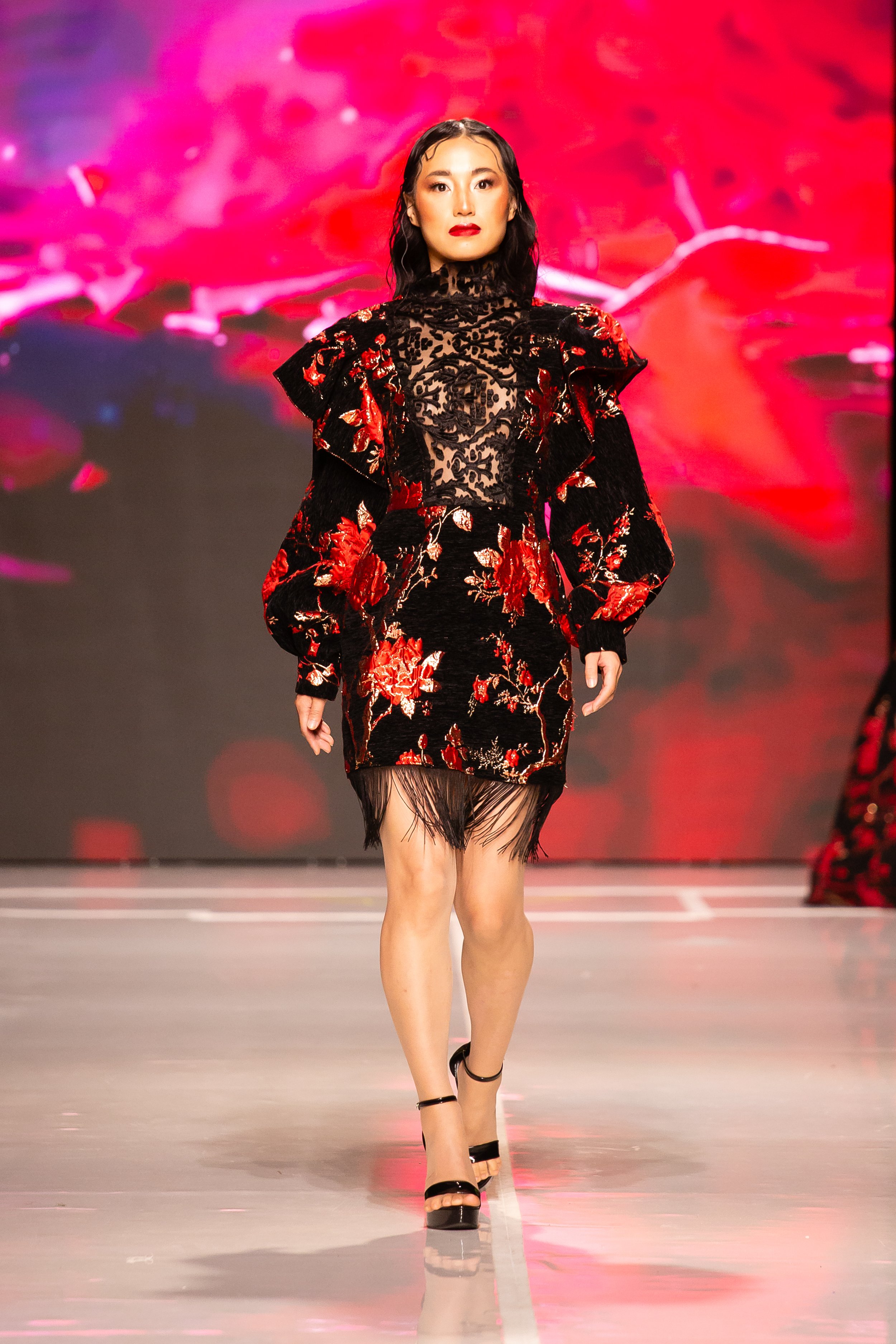 A female model walking down a runway in a black dress with red floral embroidery and sheer lace top, against a colorful pink and purple background.