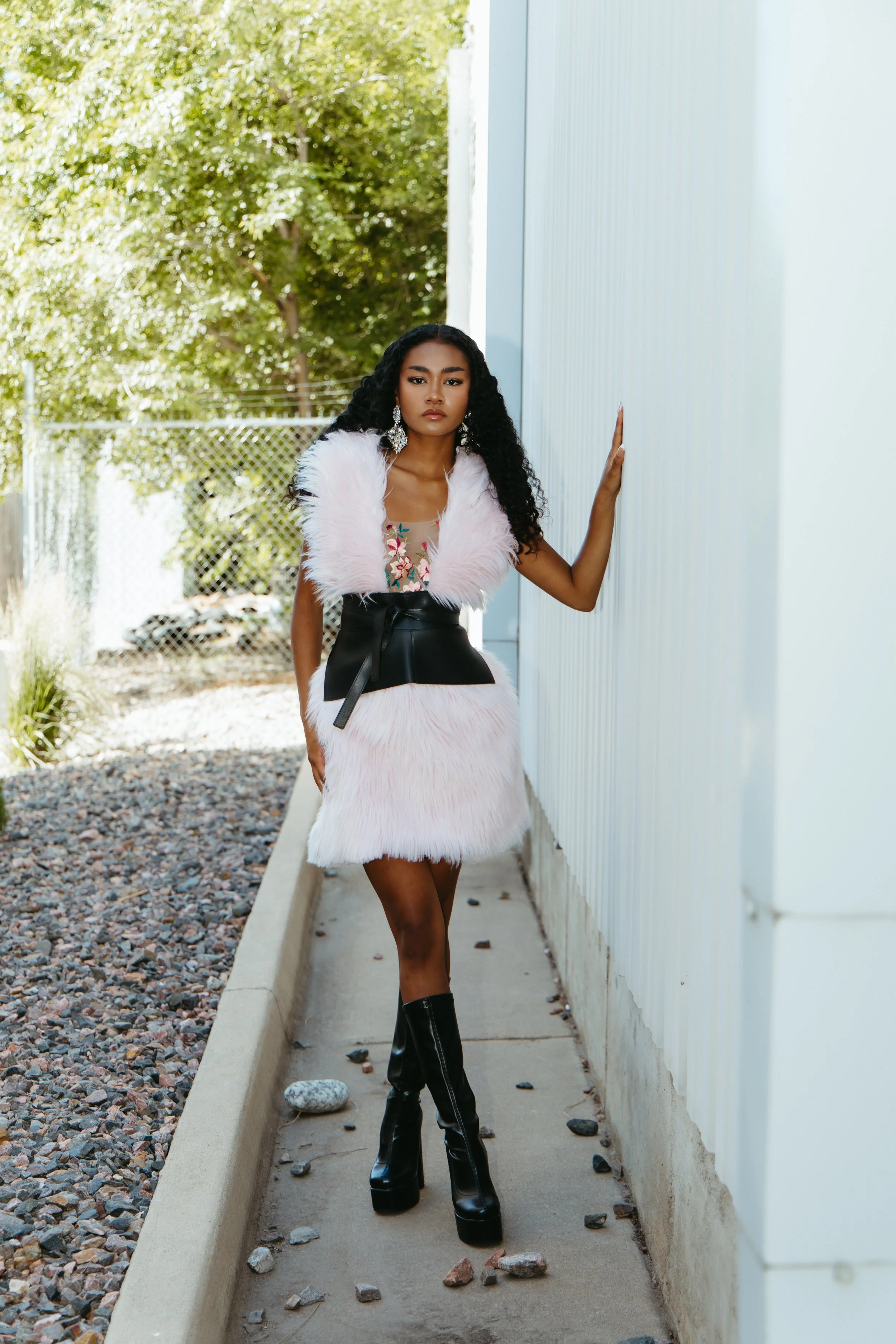A woman standing outside near a light-colored building, wearing a fashionable outfit that includes a floral top, a pink furry vest, a black leather wrap-around skirt, and tall black platform boots. She has long, curly black hair, large earrings, and a confident expression.