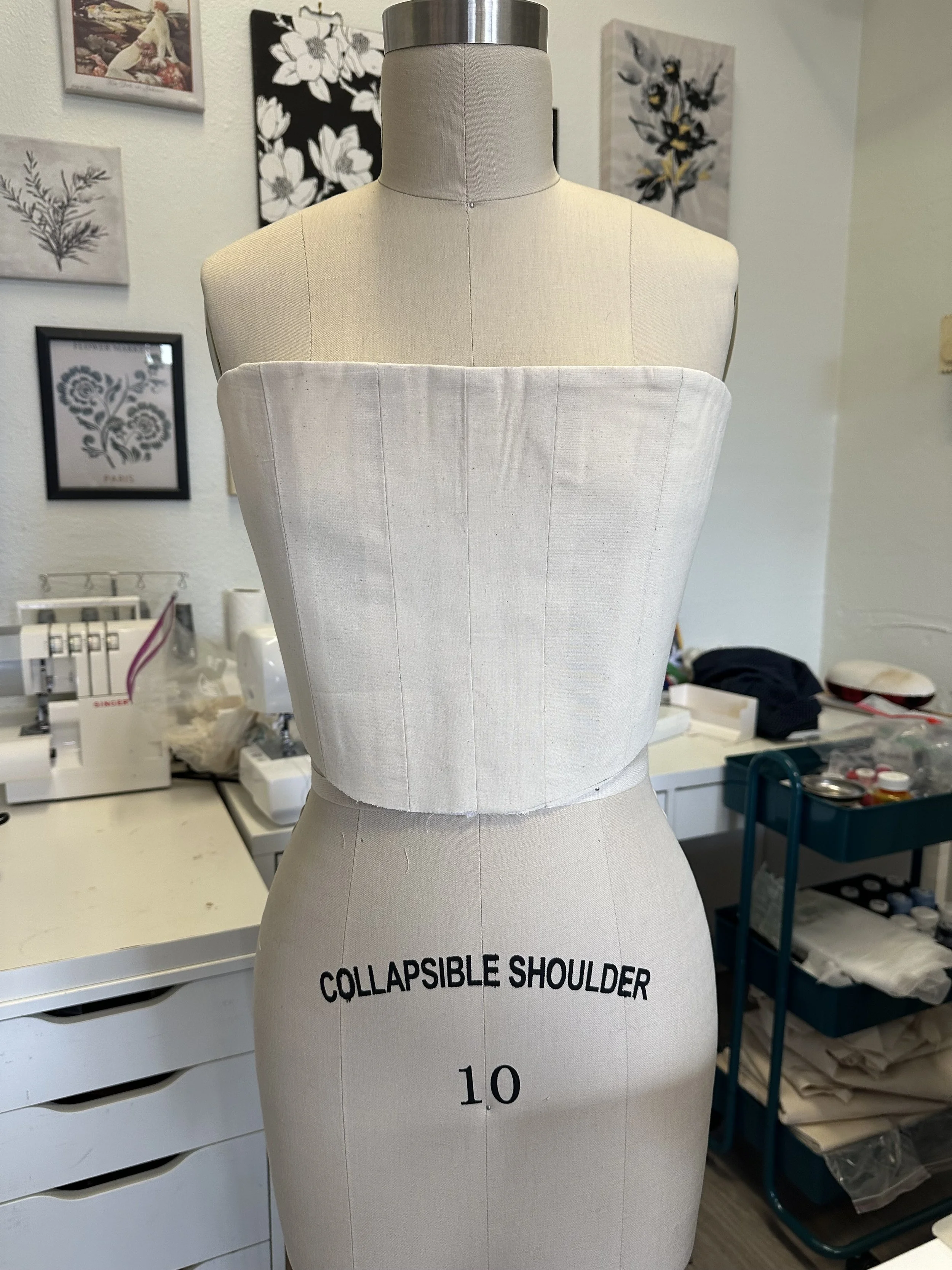 Dress form labeled "Collapsible Shoulder" in a sewing or fashion design workspace, with framed artwork and sewing supplies in the background.