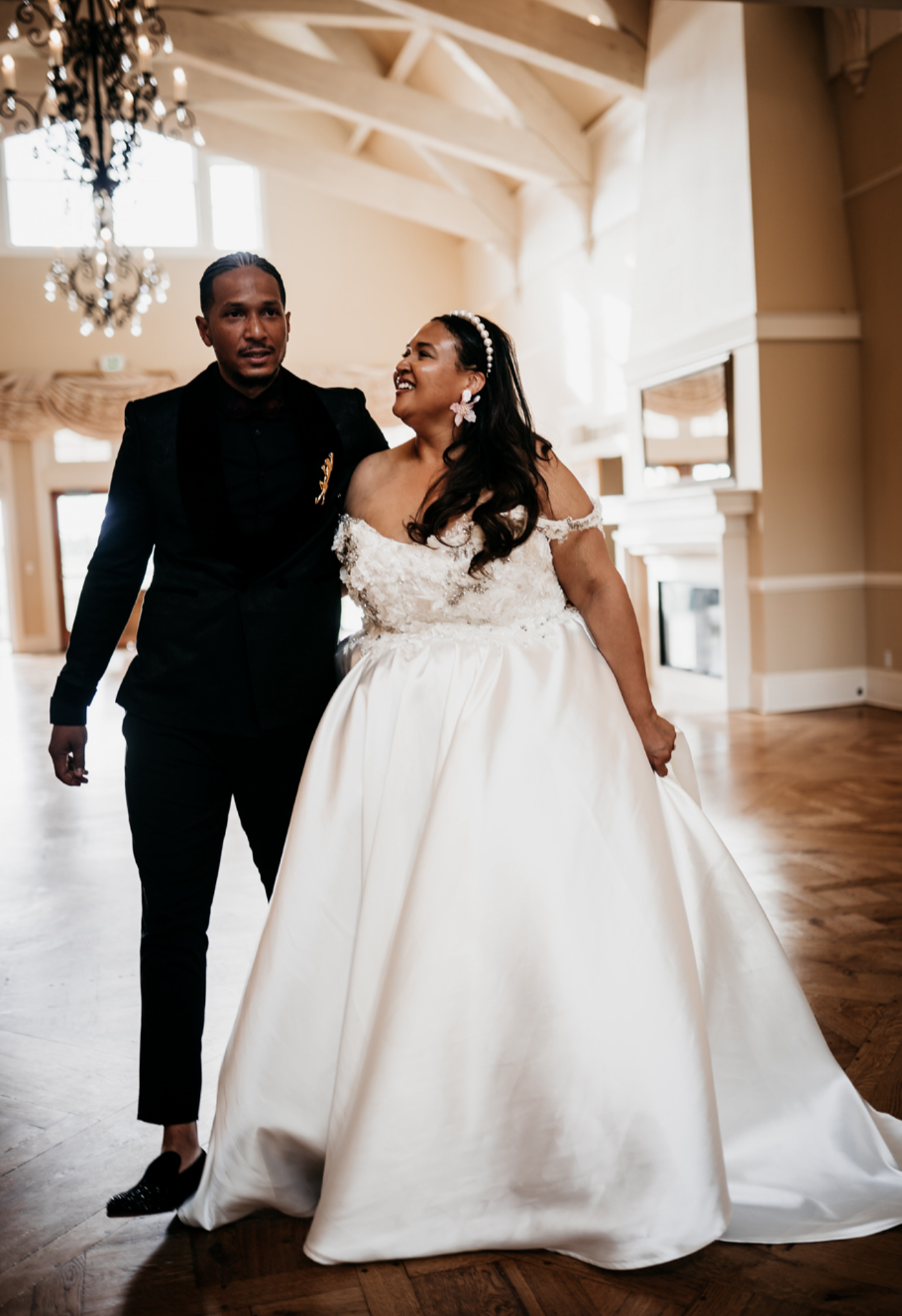 A bride in a white wedding gown and a groom in a black tuxedo dancing together in a well-lit room with wooden floors, large windows, and chandeliers.