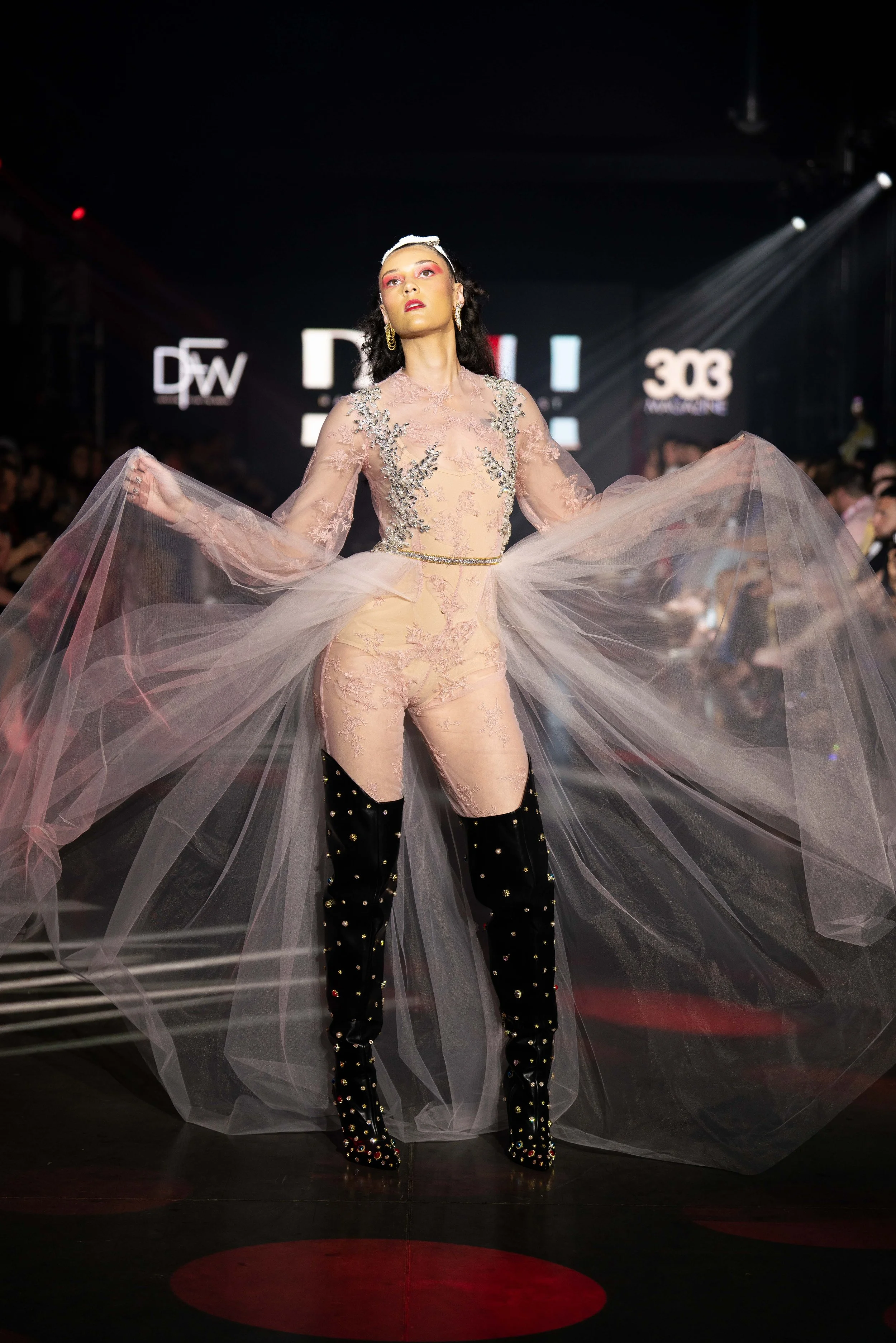 A model walking on the runway at a fashion show, wearing a nude-colored dress with floral embroidery and sheer tulle fabric, paired with black velvet thigh-high boots decorated with multicolored beads, in a dark venue with spotlights and large digita