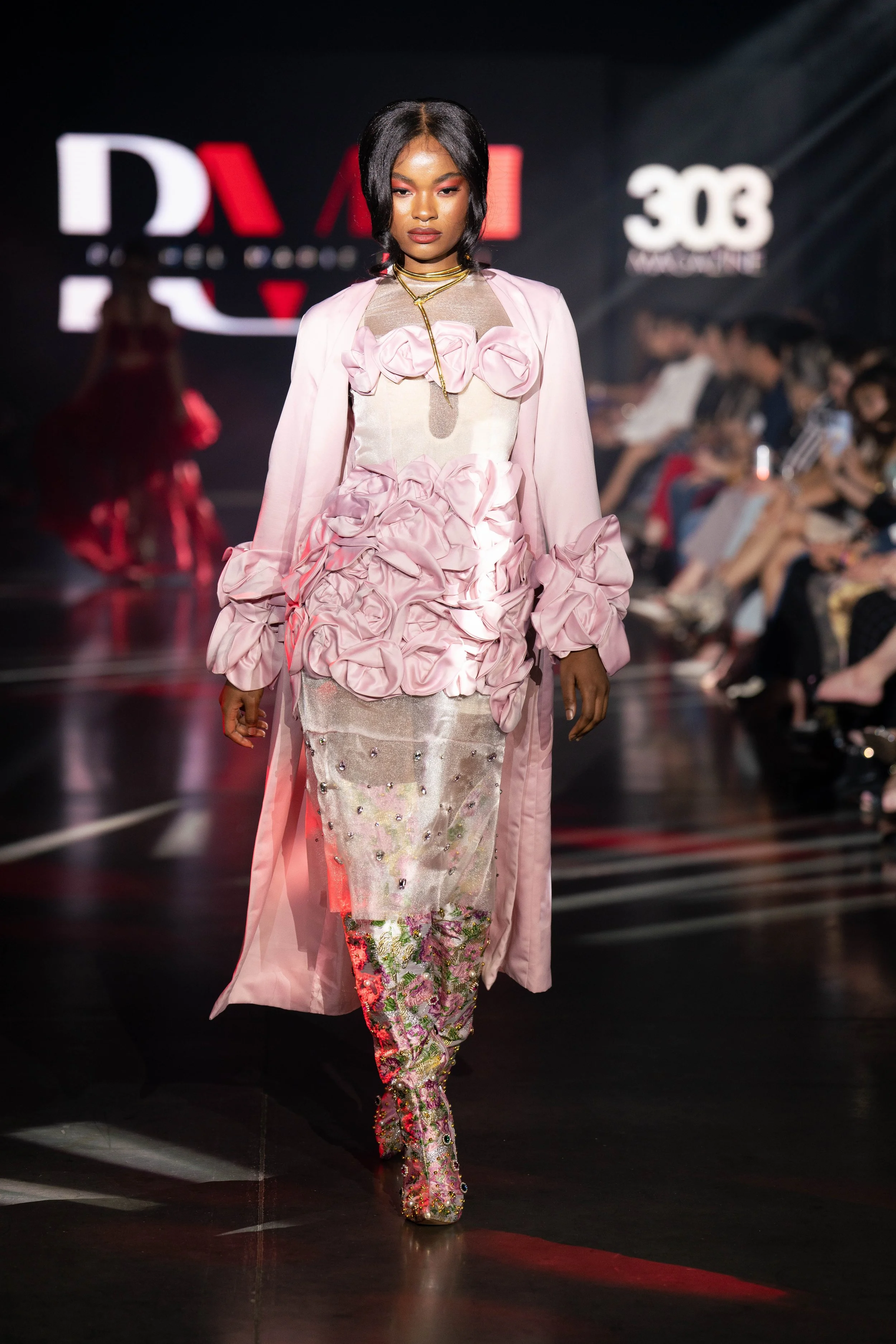 A woman walking on a runway wearing a pink dress with floral and ruffled details, paired with embroidered pants and a long pink cape, during a fashion show.