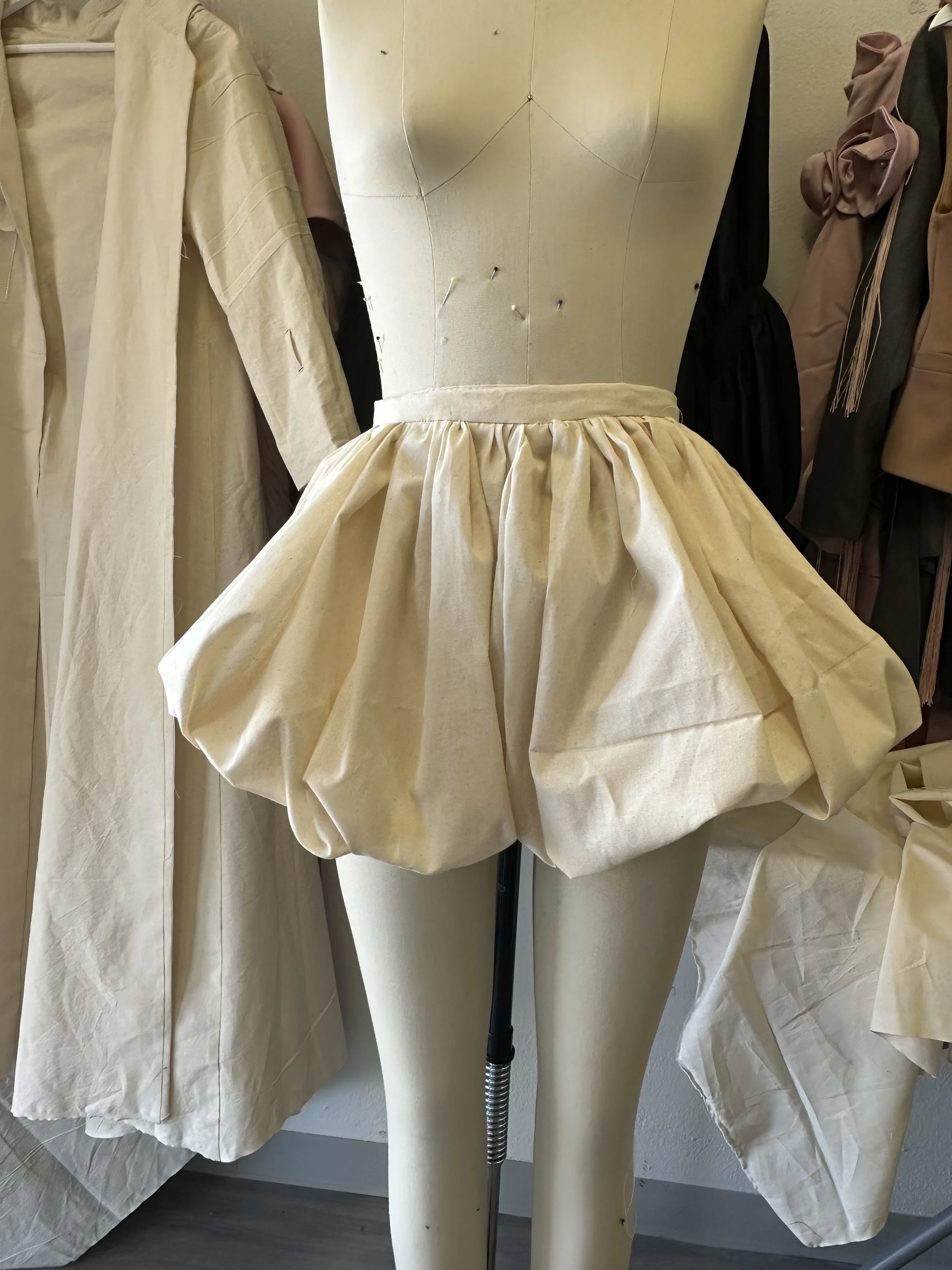 A dress form mannequin with a cream-colored skirt, surrounded by other fabric-covered mannequins and dresses in neutral tones.