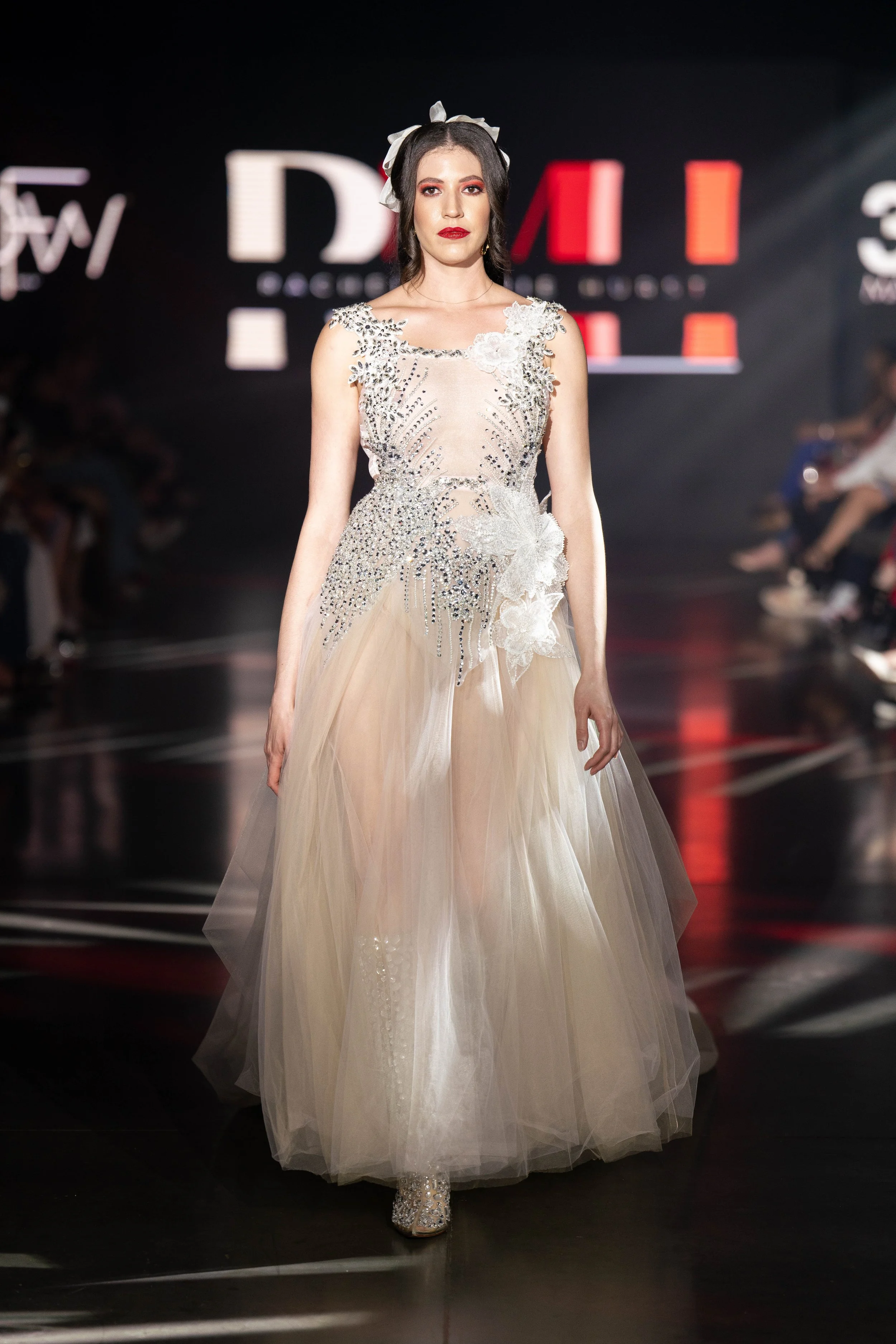 A female model walking on a runway wearing an elegant long cream dress with detailed beading and floral accents, with dark background and audience seated on either side.