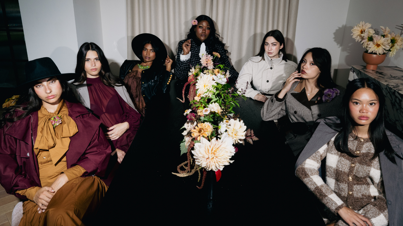 Seven women sitting around a black table with a large floral centerpiece in a well-lit room. The women are dressed in fashionable clothing, some wearing hats and styled hair, posing for the photo.