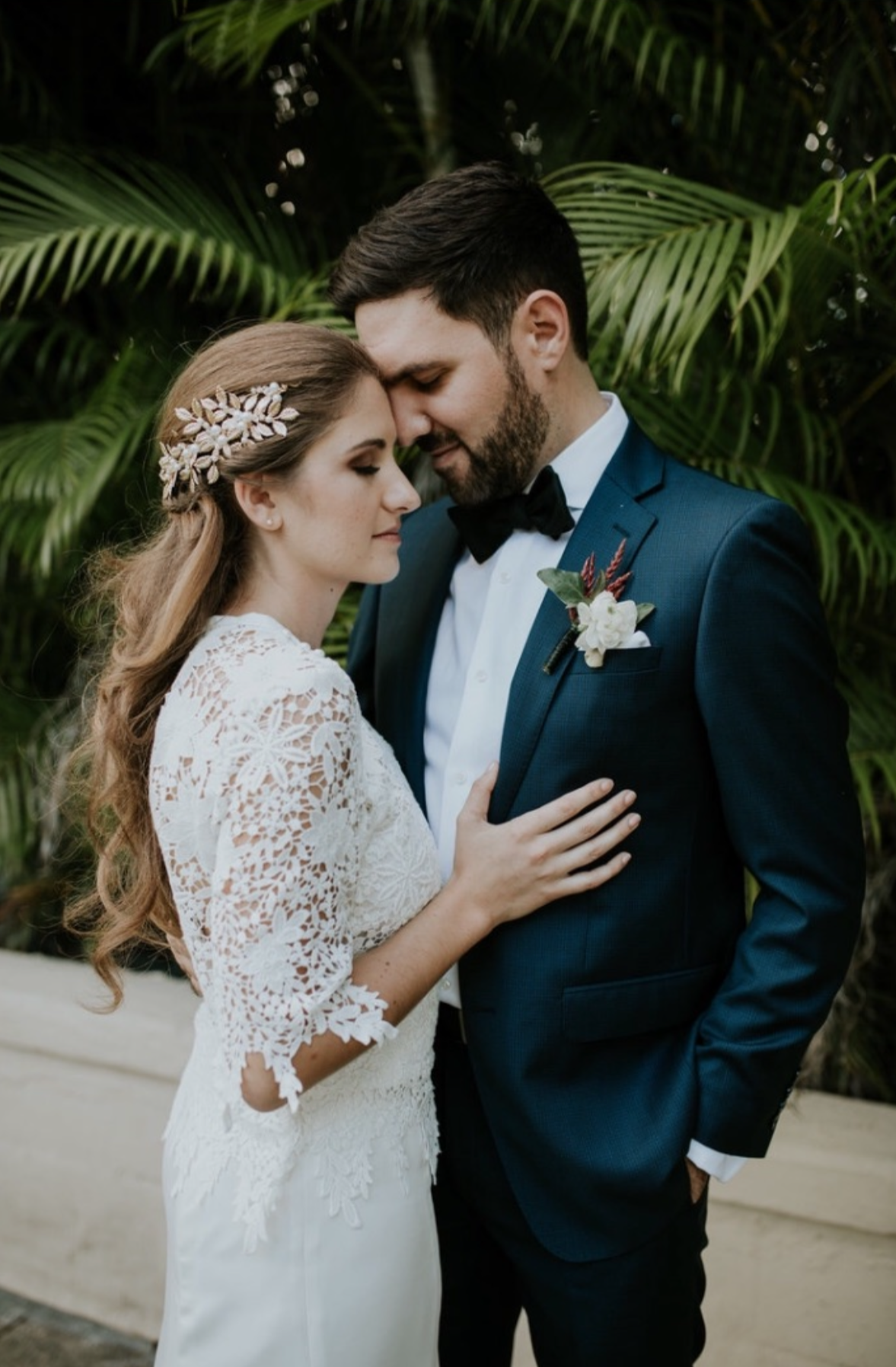 A bride and groom standing close together with foreheads touching, eyes closed, amidst lush green tropical plants, during a wedding or special occasion.