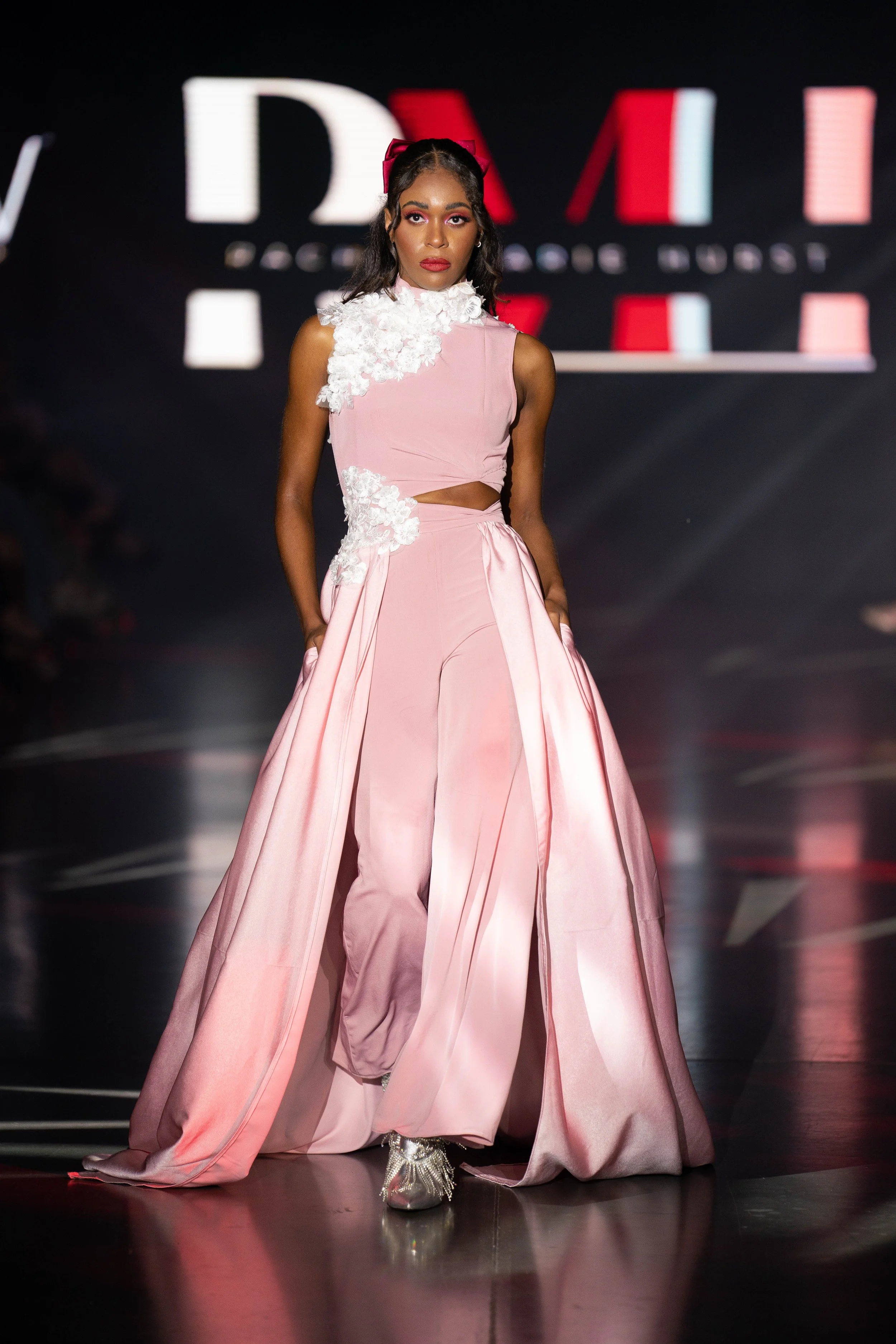Model on runway wearing a pink dress with floral accents and high boots during a fashion show.