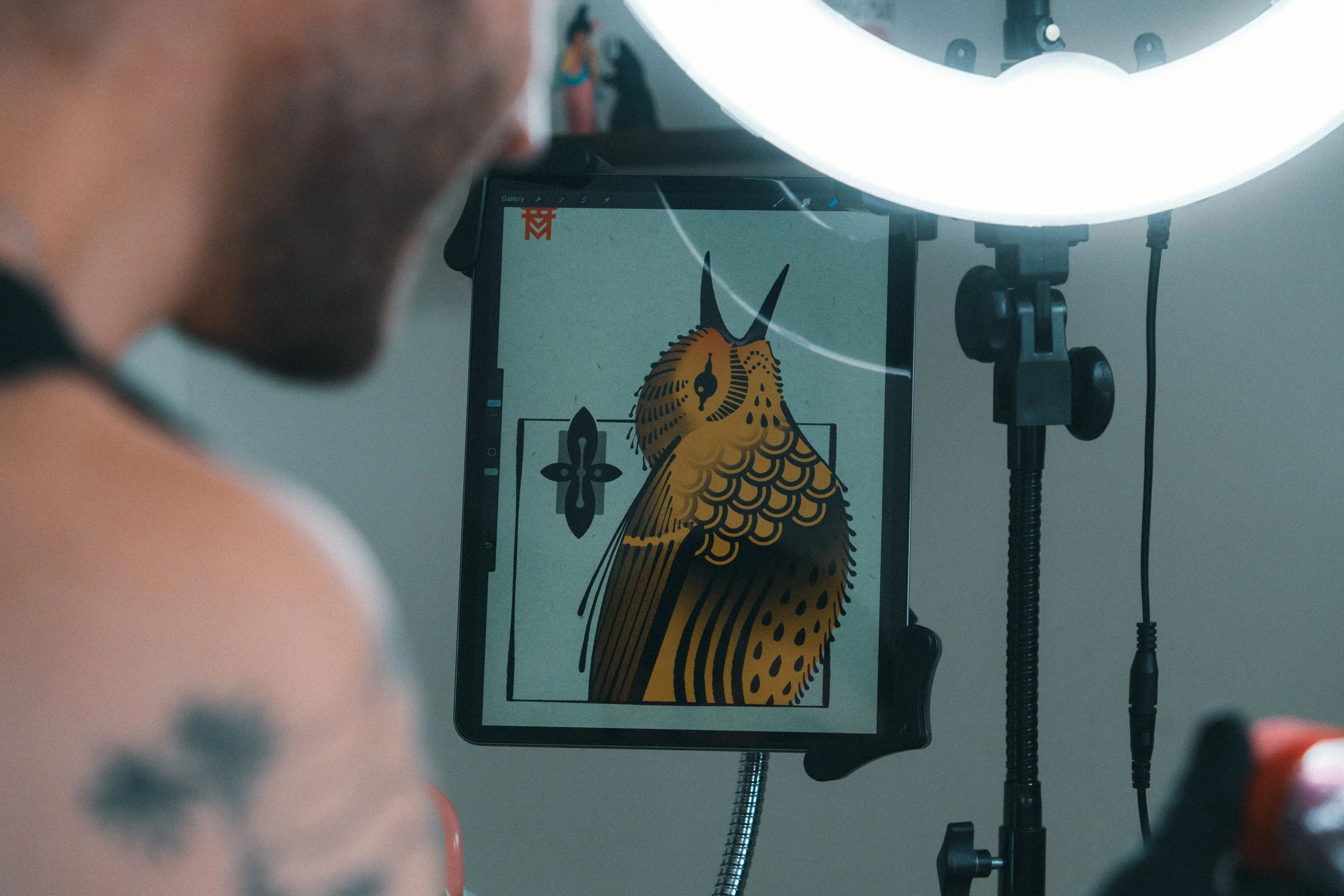 Close-up of a tattoo artist's station with a digital drawing of a stylized owl on a tablet, surrounded by a ring light, and part of a person's tattooed arm visible.