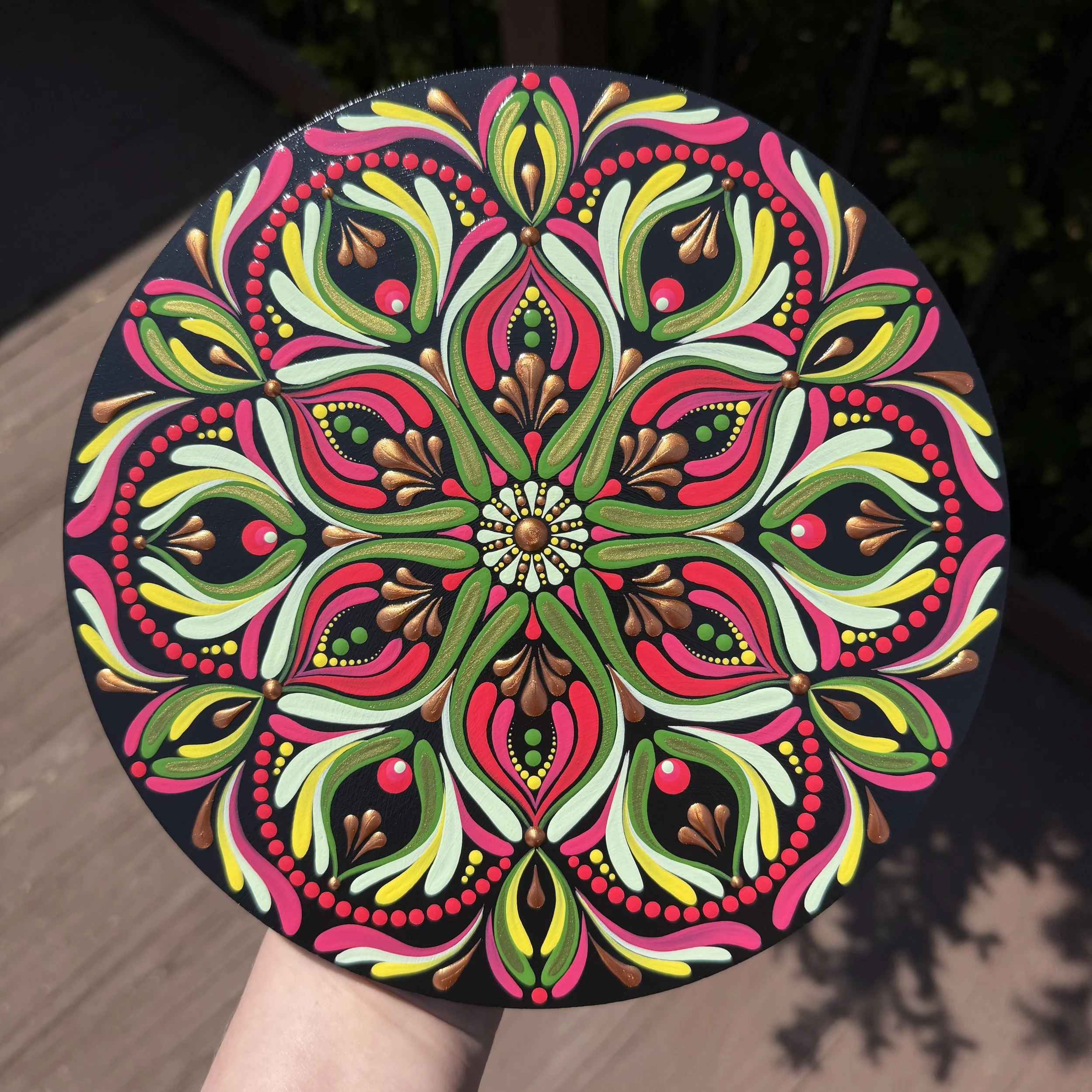 Close-up of a hand holding a round, hand-painted mandala with floral patterns in bright pink, green, yellow, white, and gold on a black background.