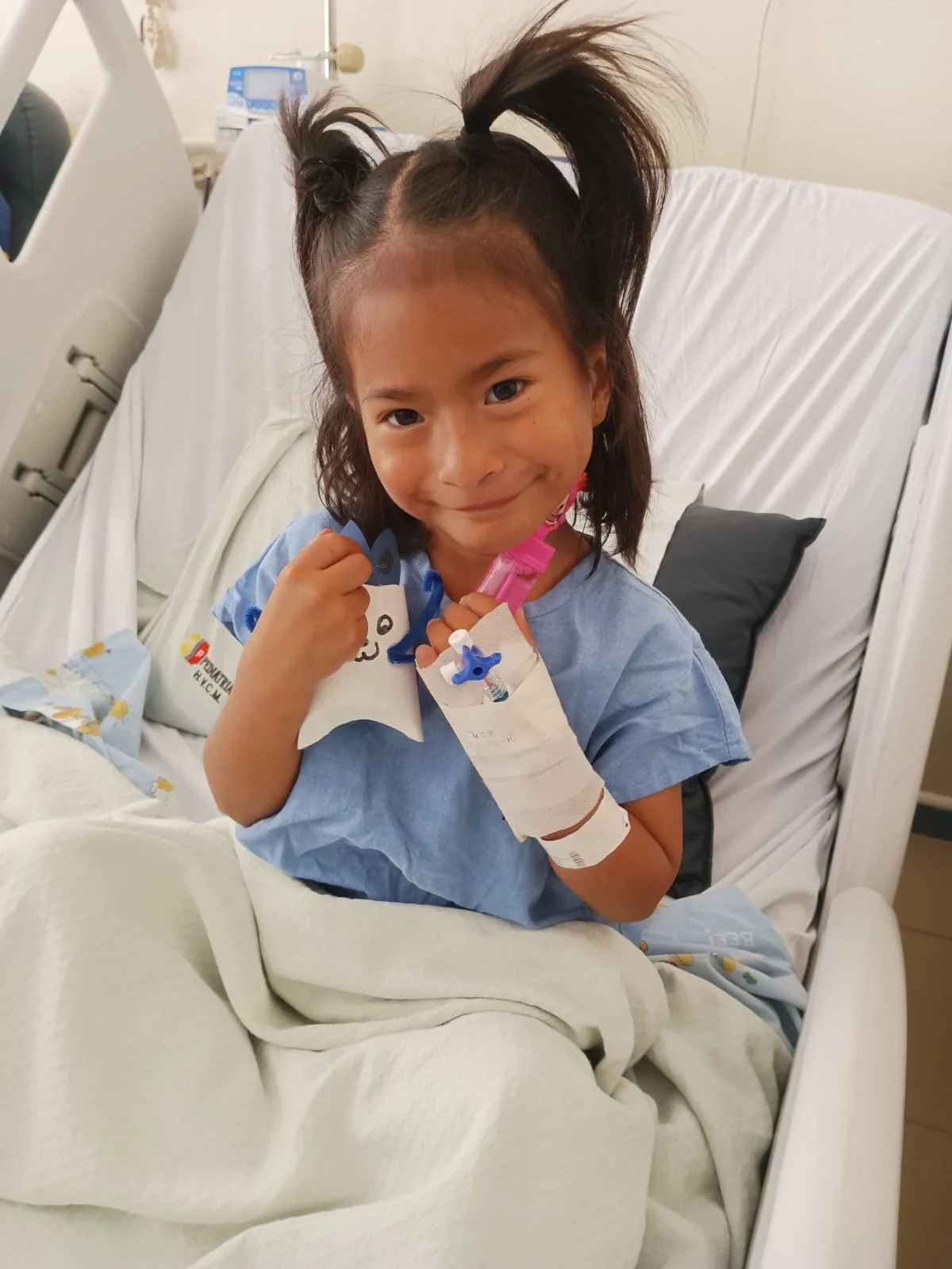 Young girl in a hospital bed smiling, holding a star-shaped card and wearing a hospital wristband and IV