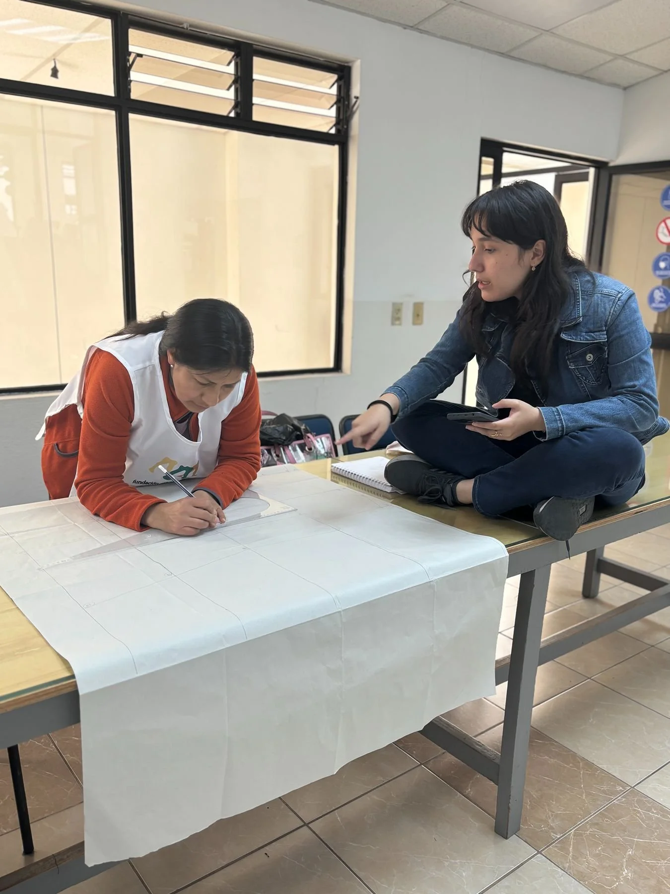 Two women engaged in a conversation at a table, with one woman sitting on a raised surface and the other leaning over the table, both focused on a drawing or project on the table.