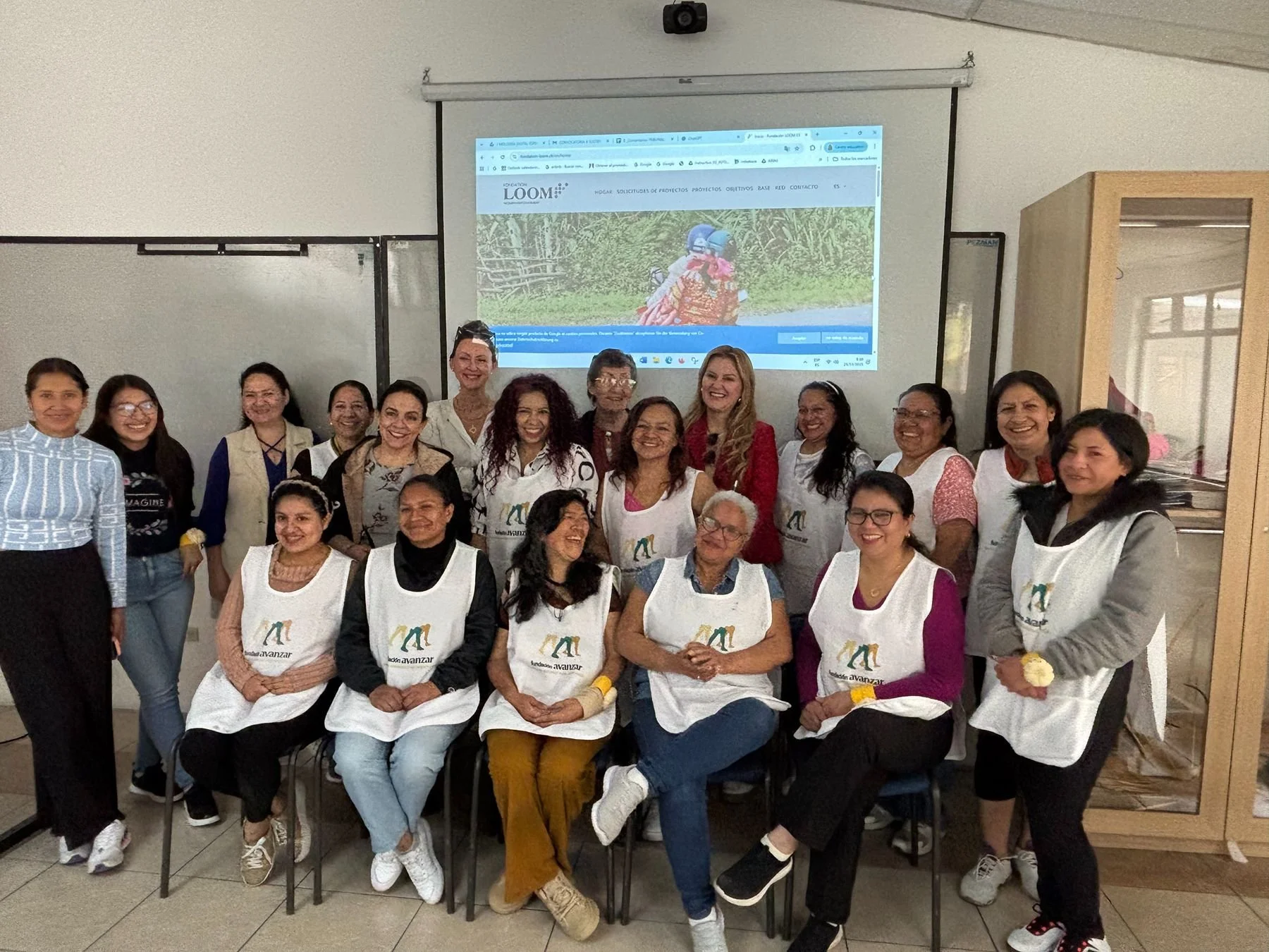 Group of women gathered in a classroom, some wearing white aprons with a logo, posing for a photo with a projected webpage in the background.