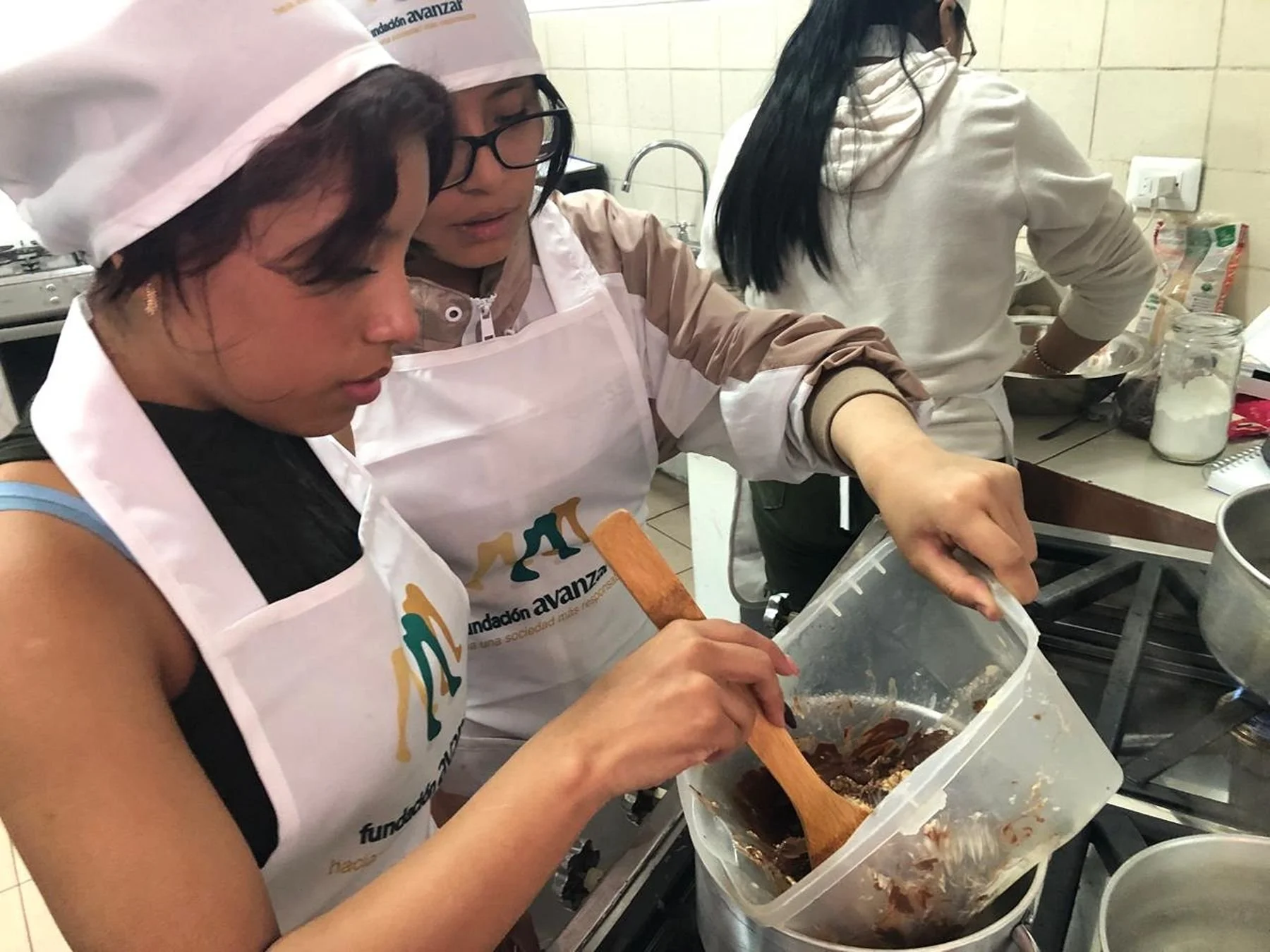 Two young women cooking in a kitchen, wearing aprons and hats, stirring a mixture in a plastic container over a stove.