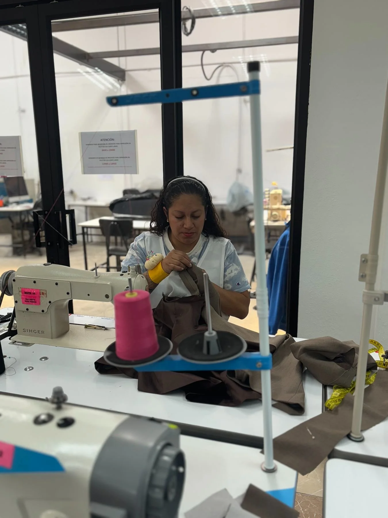 Woman working with sewing machine in a garment workshop, handling brown fabric, with spools of thread and measuring tape on the workspace.