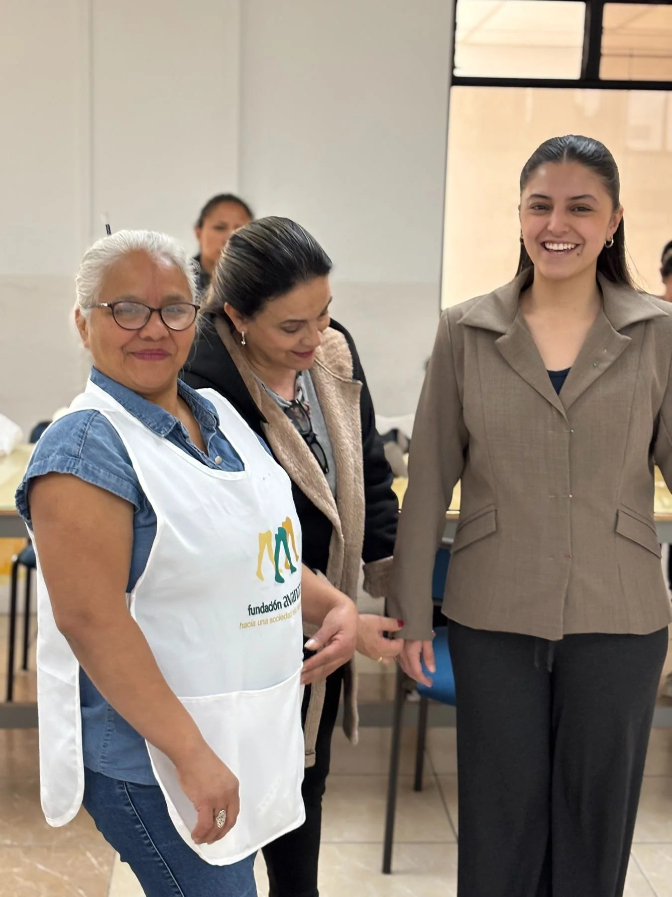 Three women standing together indoors, holding hands. The woman on the left wears glasses and a white apron with a logo, smiling at the camera. The woman in the middle is looking down, dressed in dark clothing. The woman on the right is smiling broadly, wearing a tan blazer and black pants.