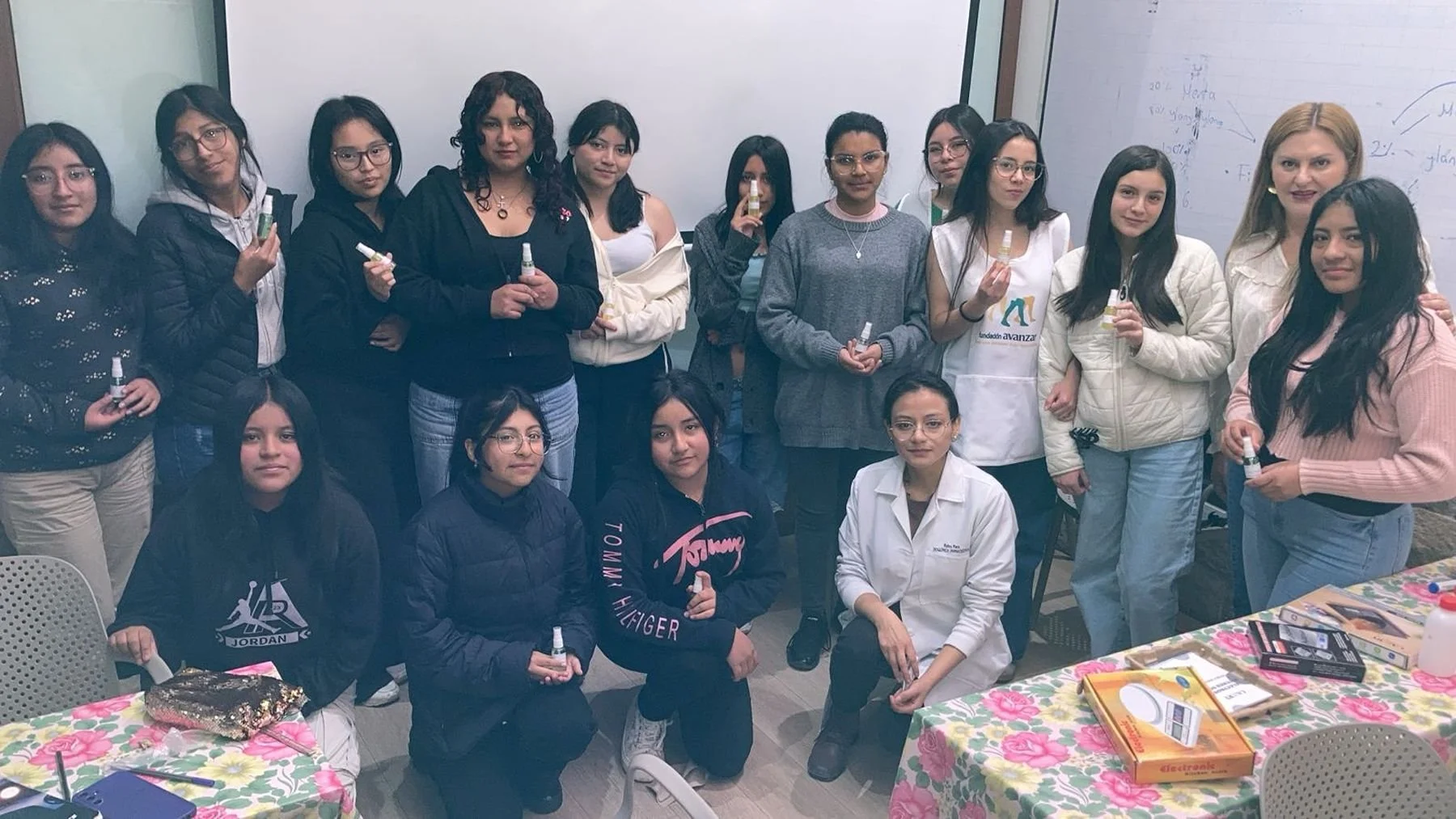 Group of young women and an instructor in a classroom, holding small bottles. Whiteboard with notes and diagrams is in the background, and tables with science kits are in the foreground.