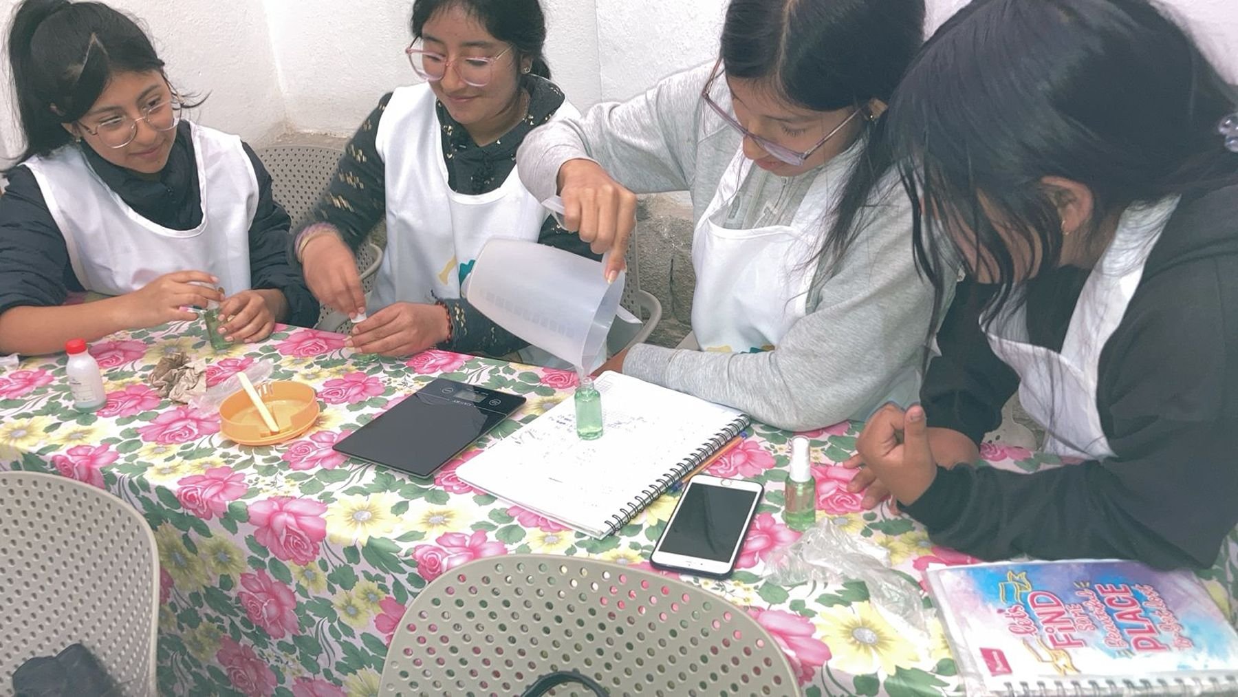 Group of women conducting a science experiment with small bottles and tools at a pink floral table.