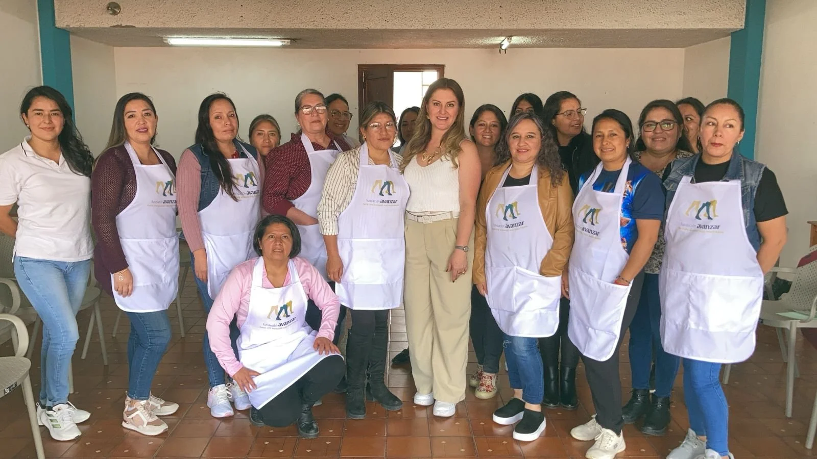 Group of women, some wearing aprons with a logo, posing together inside a room with white walls and a brown tiled floor.