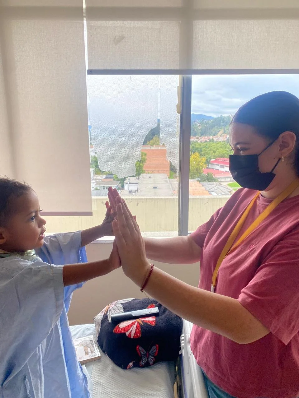 A young girl in a hospital bed giving a high five to a woman wearing a face mask, with a broken window in the background.