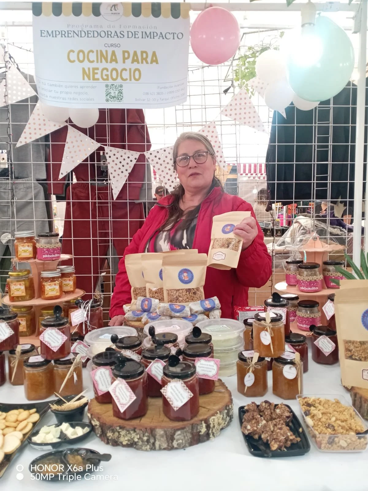 Woman at a market stall selling homemade jams, honey, and baked goods, with a sign promoting a cooking course for entrepreneurs in impact.