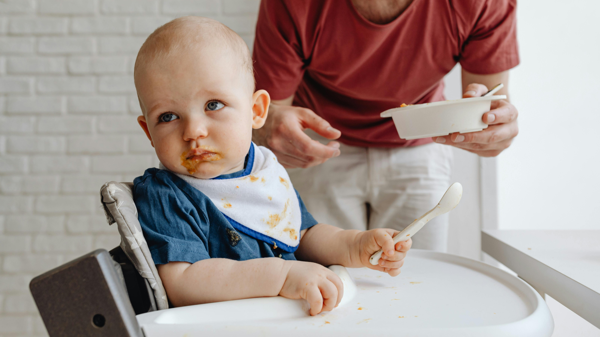 Toddler refusing food in high chair while parent tries to feed him, illustrating picky eating and feeding challenges in toddler feeding development