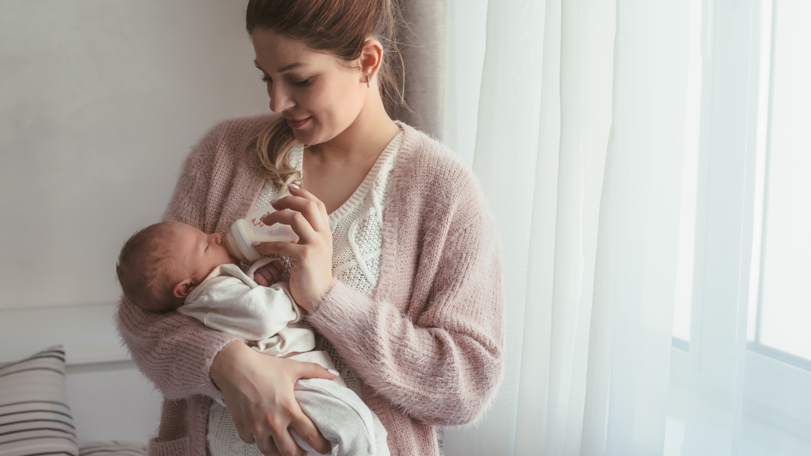 Parent bottle feeding a newborn baby by a window