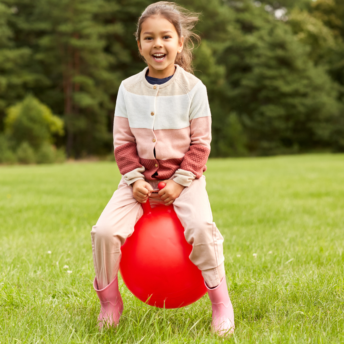 Young girl laughing while sitting on a red bouncing ball outdoors on green grass, with a wooded background.