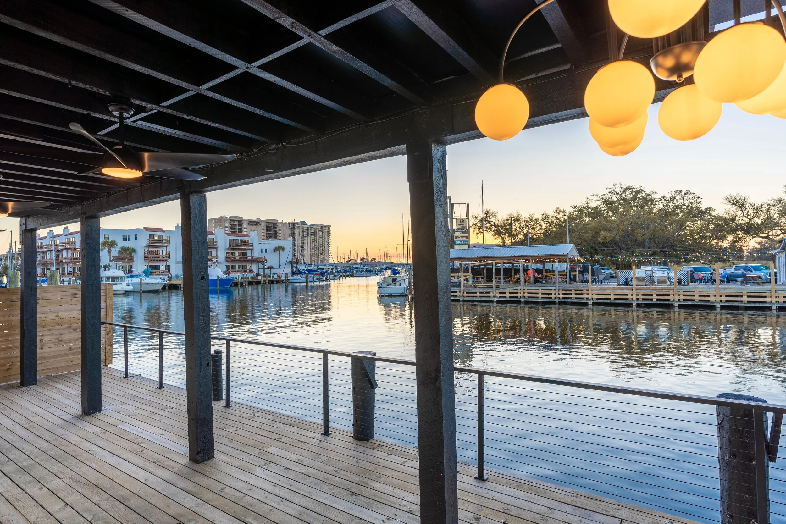 View from a covered wooden dock overlooking a marina with boats, modern buildings, and trees at sunset, with hanging round yellow lights in the foreground.
