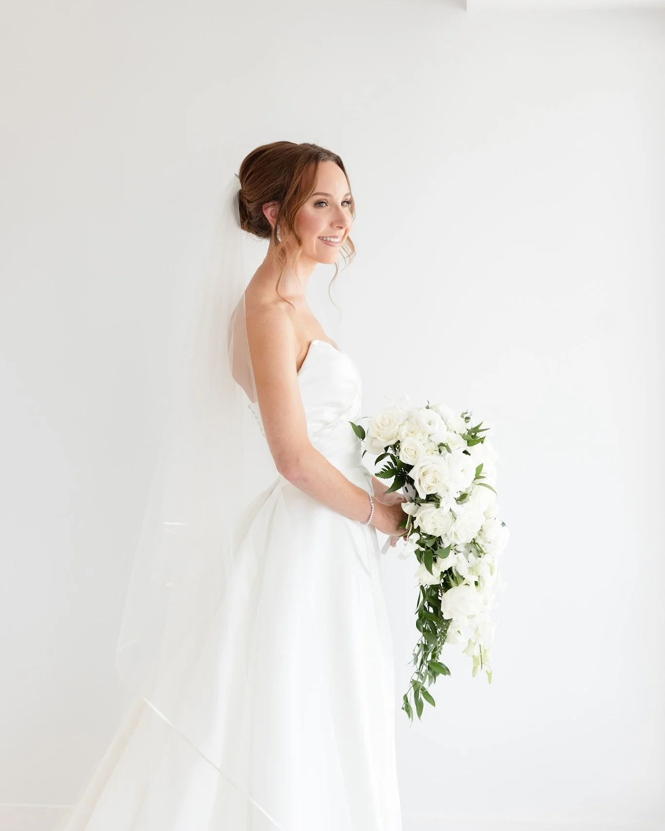 A smiling bride with brown hair styled in an updo, wearing a white satin strapless wedding gown and holding a cascading bouquet of white roses and greenery, standing against a plain white background.