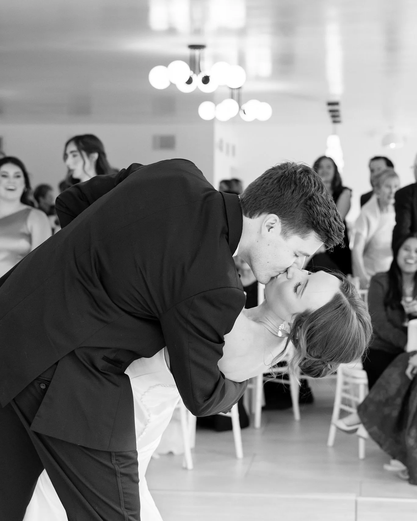 A black and white photo of a bride and groom sharing a kiss at their wedding reception, with guests watching and smiling in the background.