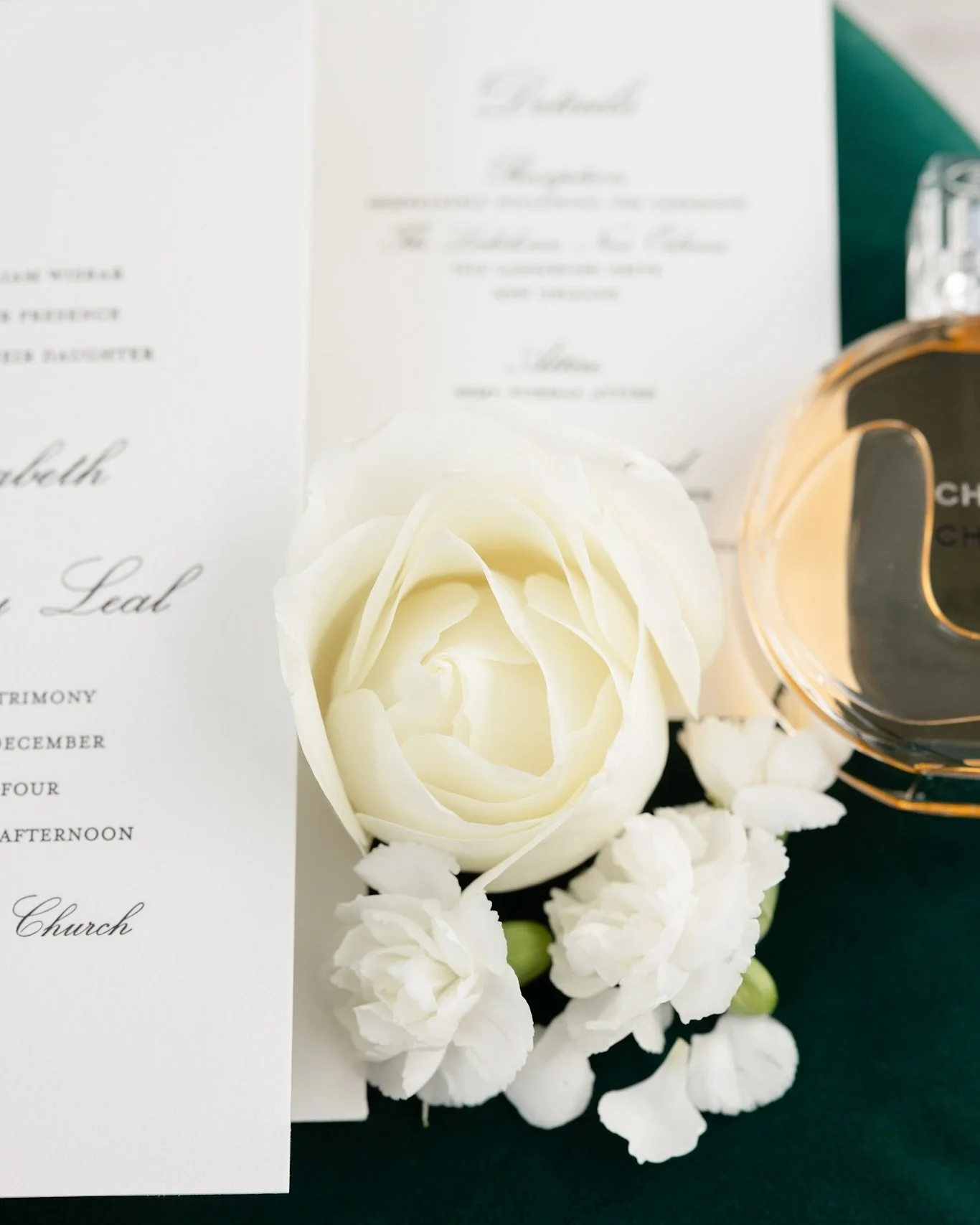White rose surrounded by white flowers, a partially visible perfume bottle, and wedding invitations on a green surface.